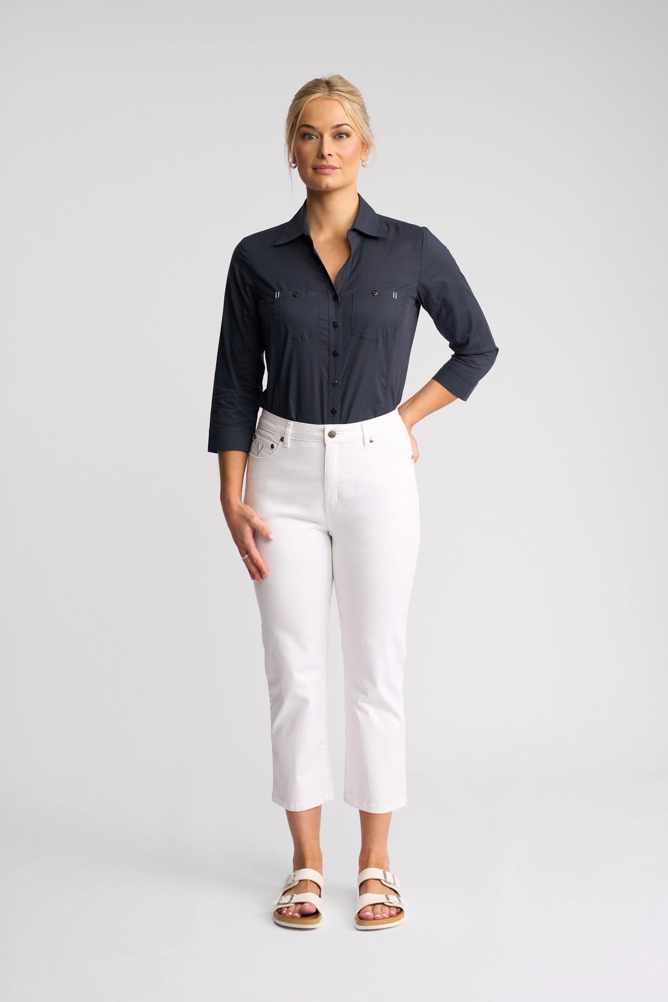 A woman stands facing forward, wearing a navy button-up shirt paired with Vassalli’s Arrival Pant in white—crafted from comfortable stretch cotton in a crisp 7/8 length slim-leg silhouette. She completes her outfit with white sandals. Her blonde hair is tied back, and she stands against a plain, light background.
