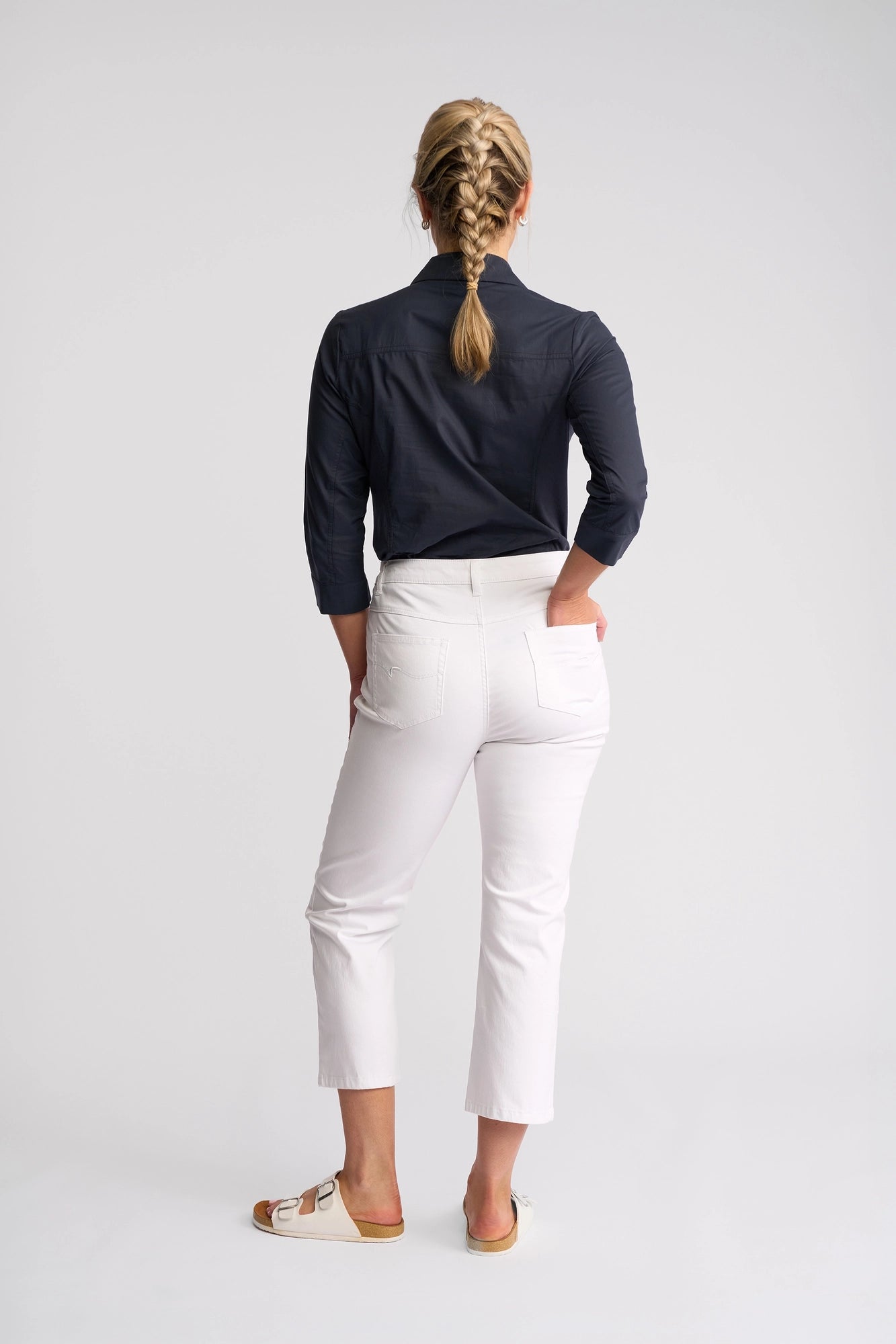 A woman stands facing away, dressed in a black long-sleeve shirt and the Vassalli Arrival Pant - White, featuring a 7/8 length slim-leg silhouette. She pairs the stretch cotton pants with white sandals, her blond hair styled in a braid as she places her left hand in the pocket. The plain, light background draws attention to the sleek design of these Vassalli pants.