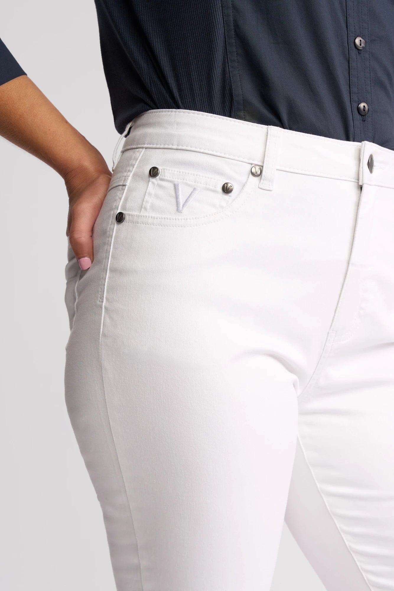 A person wearing a navy blue shirt and Vassalli Arrival Pant - White, showcasing the 7/8 length slim-leg style with a signature V logo on the pocket, poses confidently with one hand in their pocket.