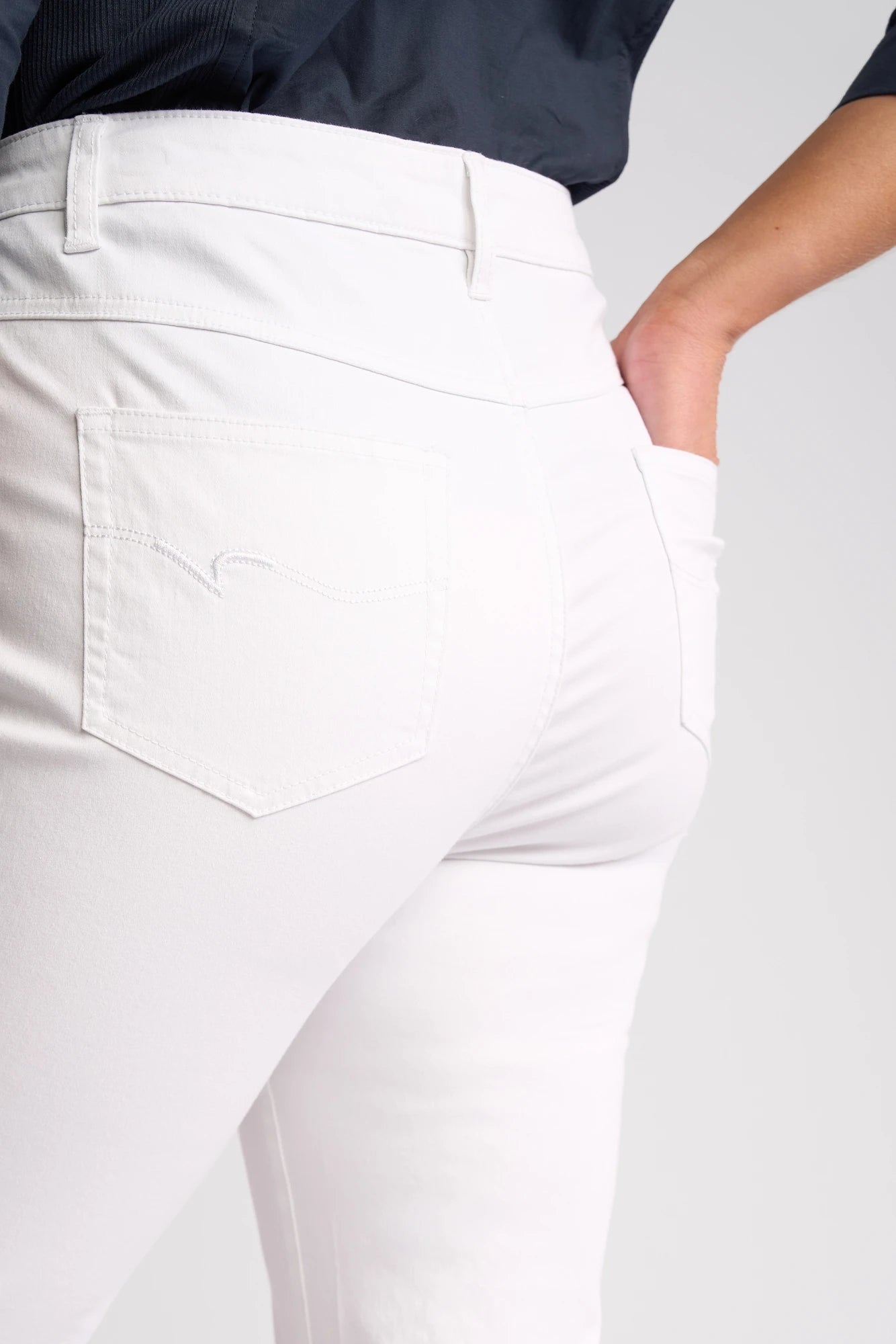 Close-up of a person wearing the Vassalli Arrival Pant - White, featuring 7/8 length slim-leg design with their right hand in the back pocket, showcasing the pocket detail and seam stitching. Crafted from stretch cotton, these pants are paired with a dark-colored top.