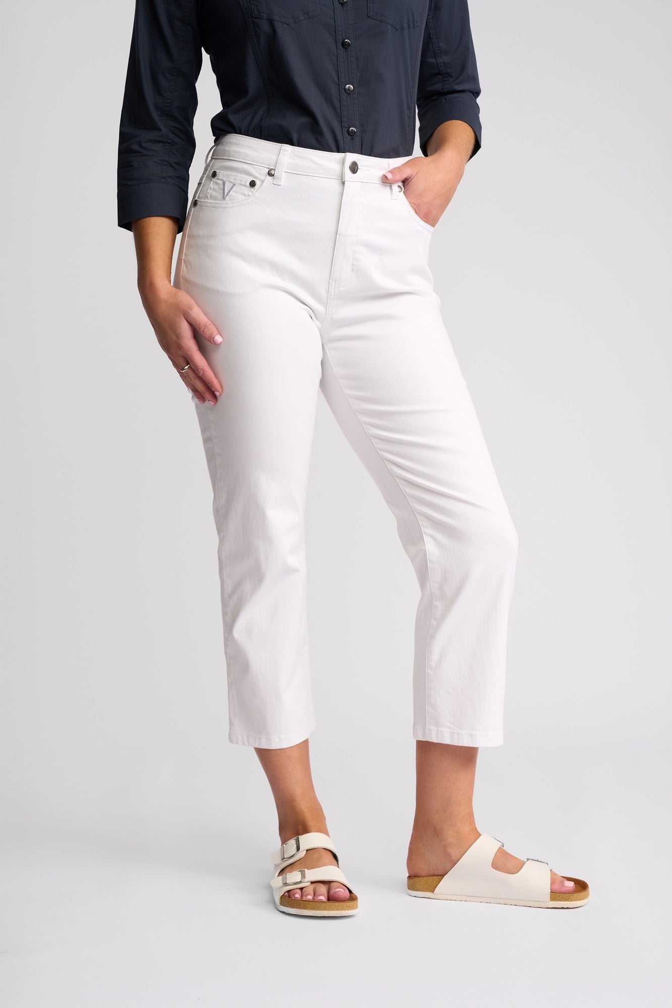 A person wearing a dark long-sleeve shirt, Vassalli Arrival Pant - White, and light-colored sandals stands with one hand in their pocket against a plain background.