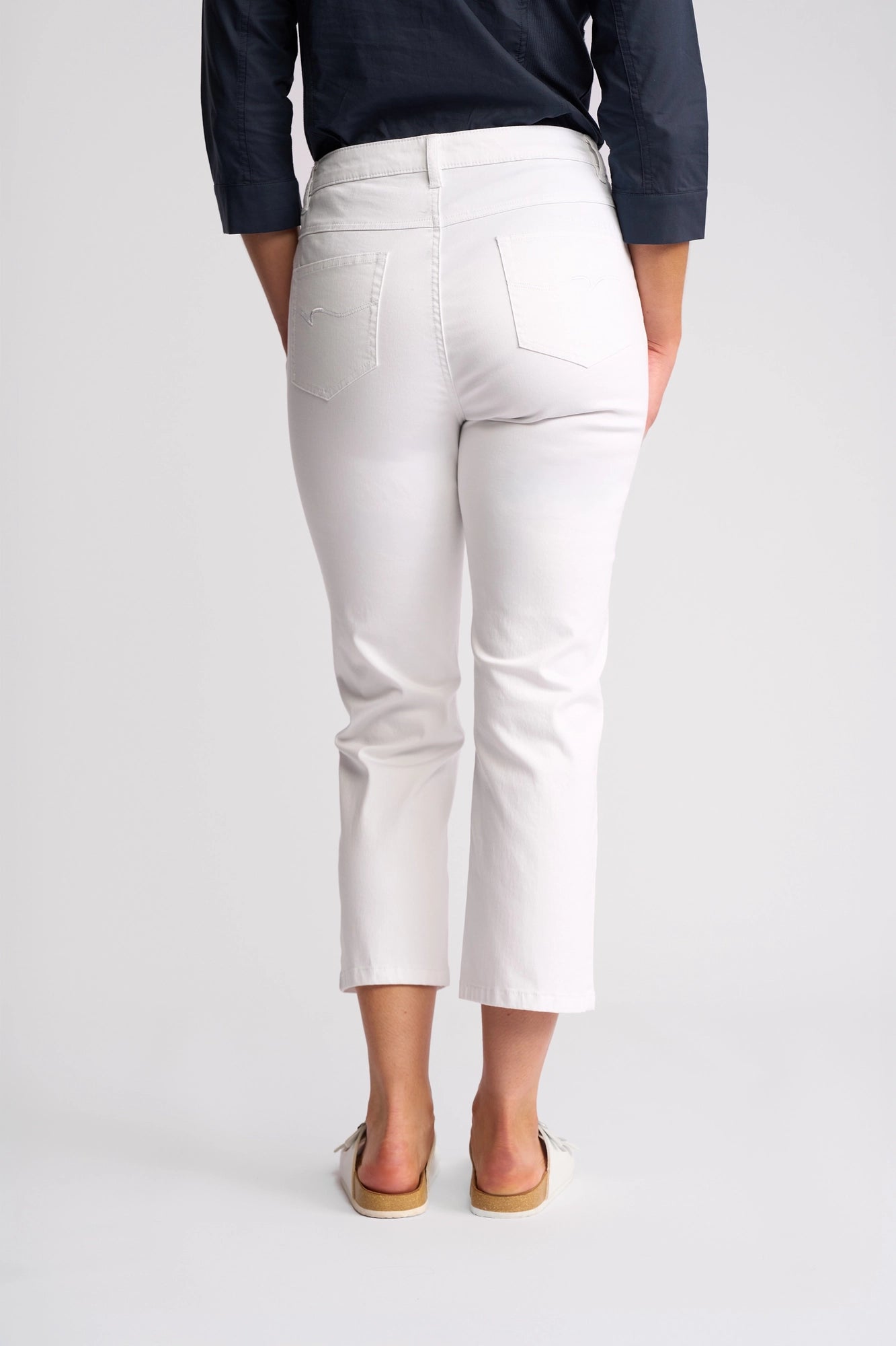 A person is shown from the back, standing barefoot on a white background, wearing the Vassalli Arrival Pant - White—a pair of 7/8 length slim-leg stretch cotton pants—paired with a dark long-sleeve shirt and sandals.