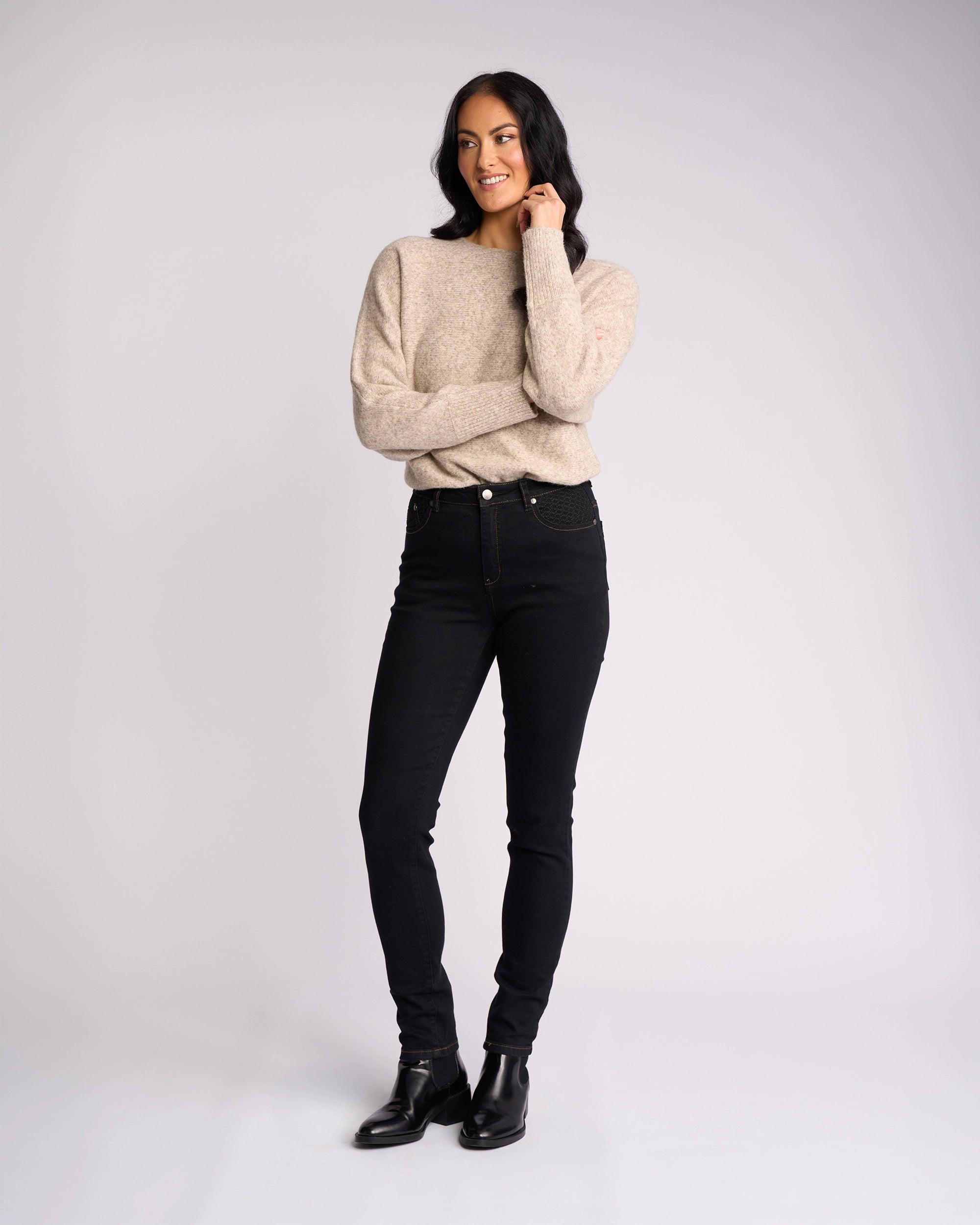 A woman stands in front of a plain backdrop, wearing the Vassalli Reign Jean - Black Denim with a beige sweater and black ankle boots, smiling and looking to the side—perfect for effortless women’s denim NZ style.