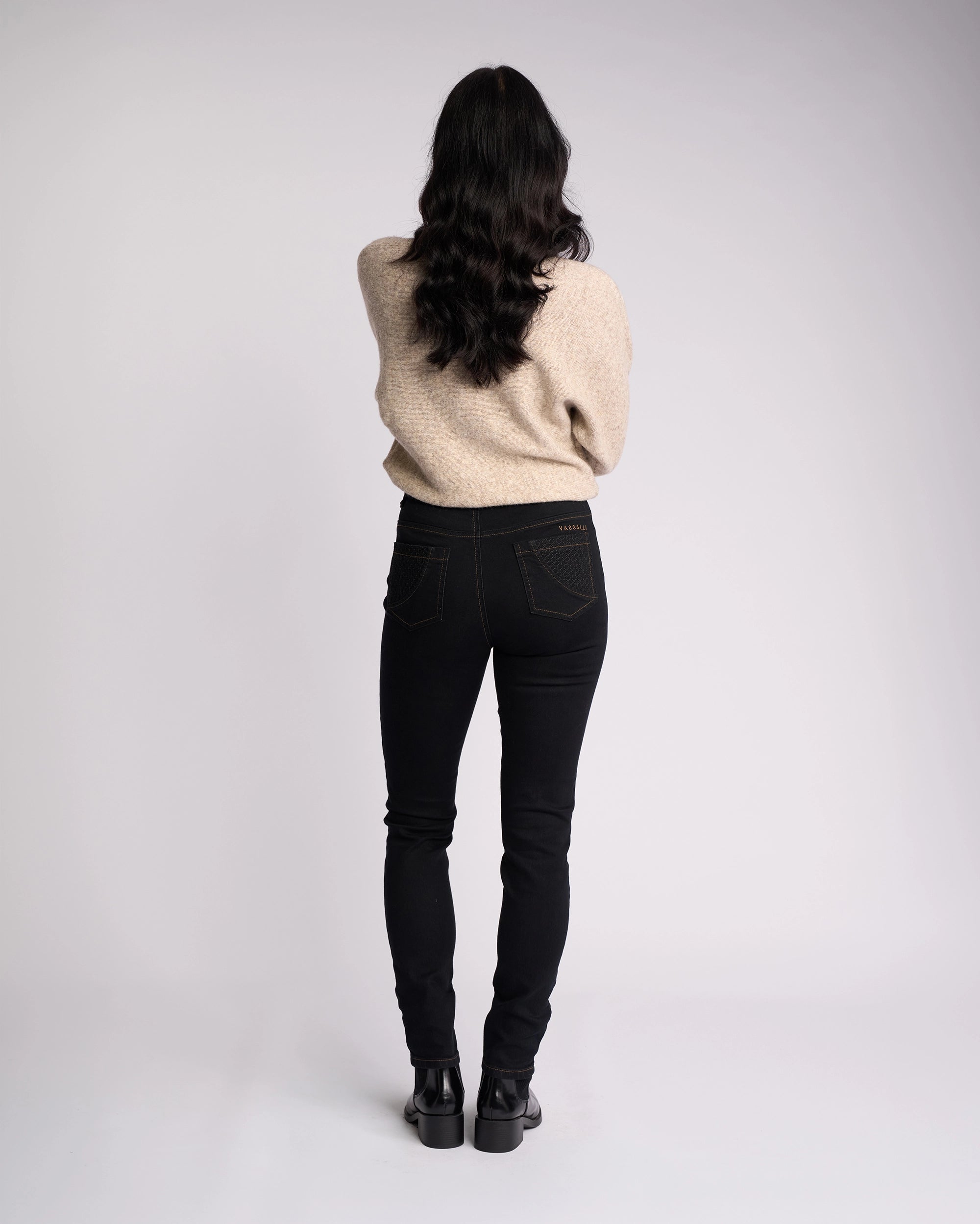 A person with long dark wavy hair stands facing away from the camera against a plain white background, wearing a beige sweater, black shoes, and Vassalli Reign Jean in Black Denim.