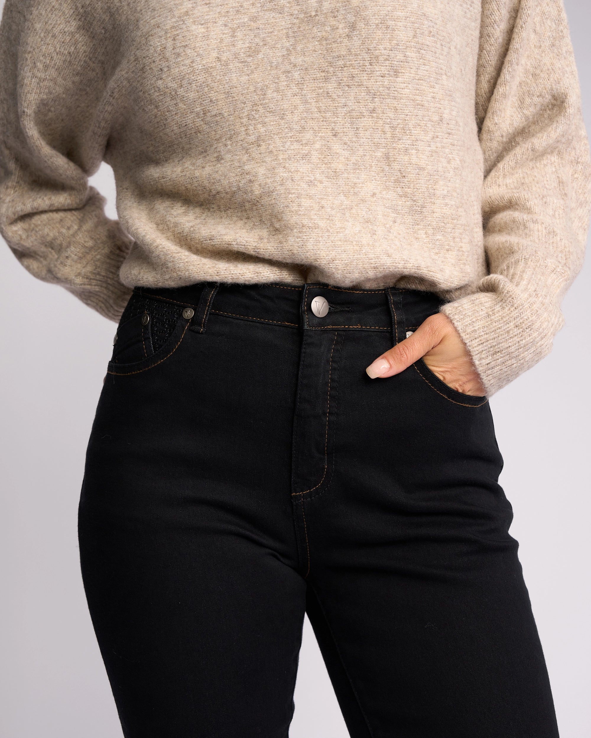 A person wearing a beige sweater and Vassalli’s Reign Jean - Black Denim stands with one hand in their pocket against a plain background, highlighting a chic women’s denim NZ style.