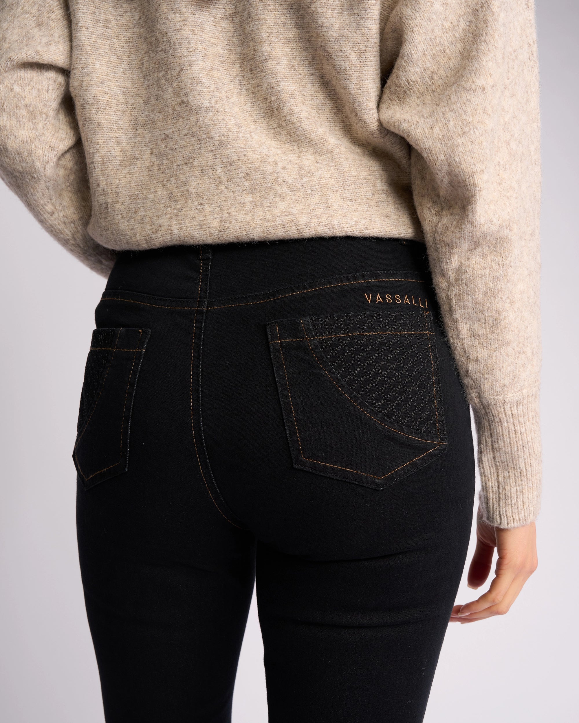 A person wearing a beige sweater and Vassalli Reign Jean - Black Denim, featuring high-waisted skinny legs, contrast stitching, and VASSALLI embroidered above the right back pocket—perfect for women’s denim NZ style.