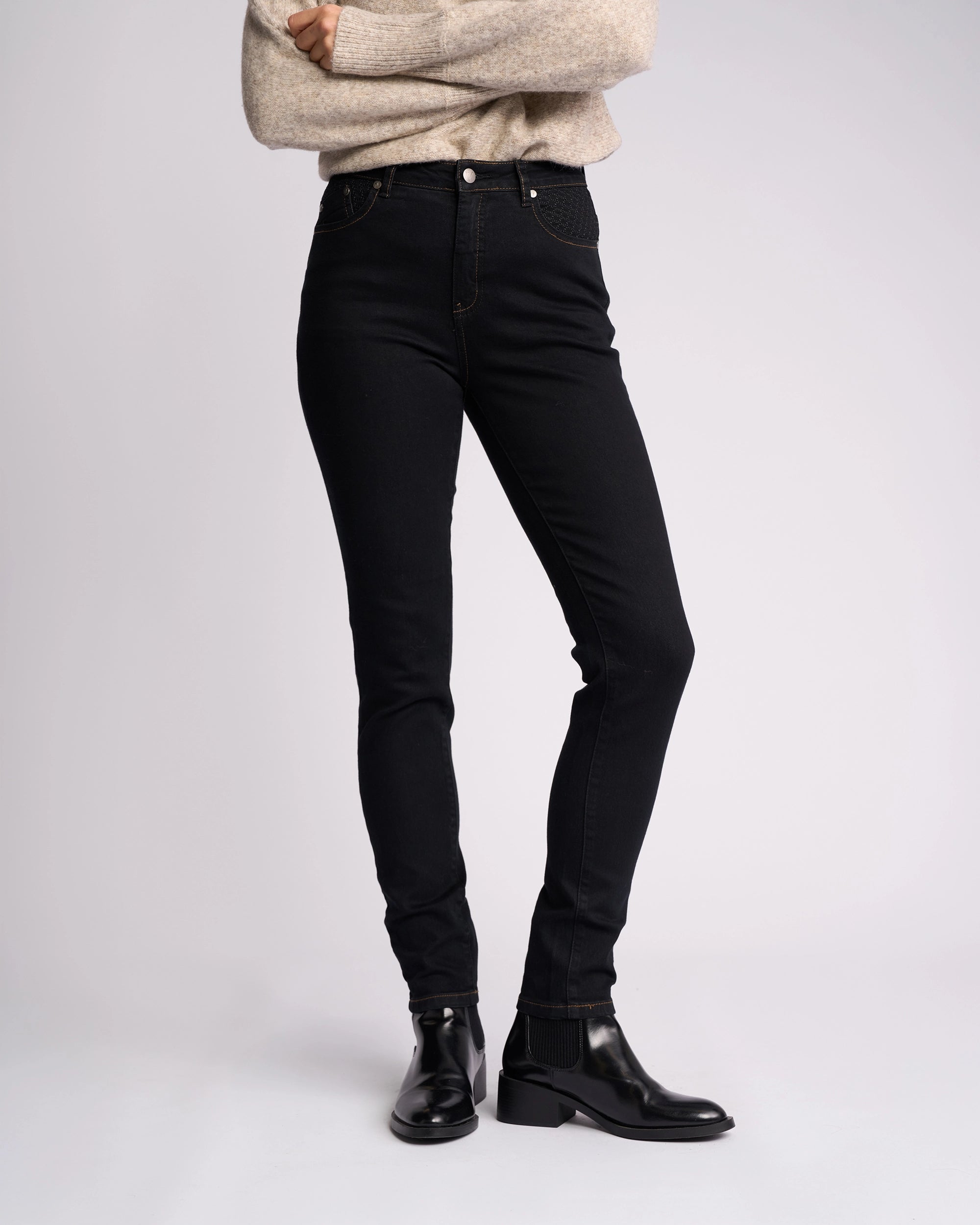 A person stands with arms crossed, wearing a beige sweater, Vassalli Reign Jean - Black Denim, and black ankle boots against a plain light background.