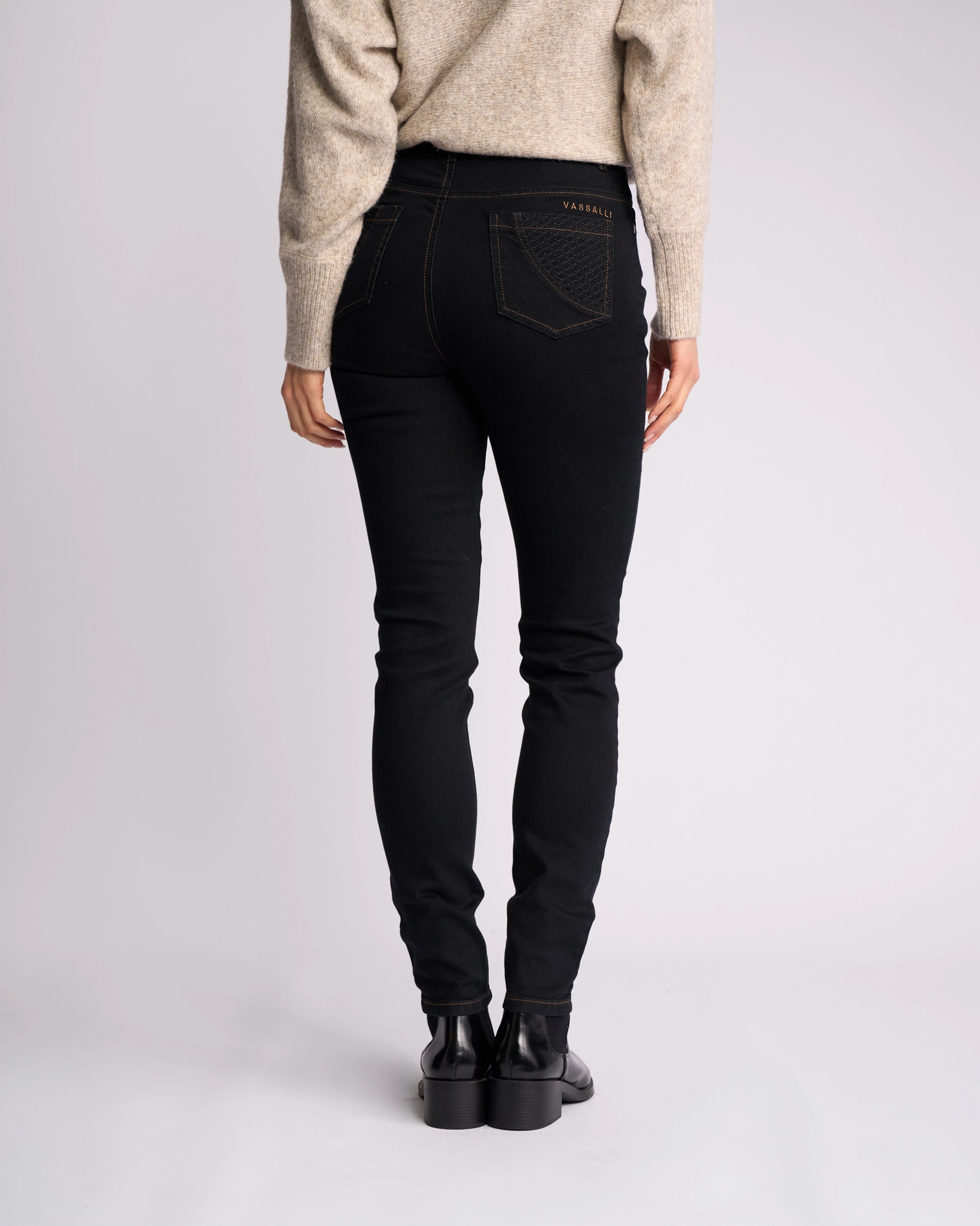 Person stands with their back to the camera, wearing a beige sweater, black boots, and Vassalli’s Reign Jean in Black Denim. Only the lower half is visible against a plain light background—a stylish pick from women's denim NZ collection.