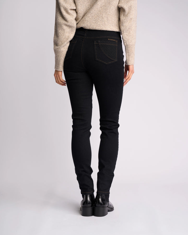 Person stands with their back to the camera, wearing a beige sweater, black boots, and Vassalli’s Reign Jean in Black Denim. Only the lower half is visible against a plain light background—a stylish pick from women's denim NZ collection.