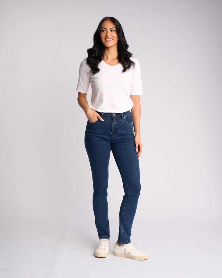 A woman stands against a plain light background, smiling in a white short-sleeve top, Vassalli Reign Jean - Brushed Indigo and white sneakers. With one hand in her pocket, she showcases effortless style in women’s denim NZ and loose waves.