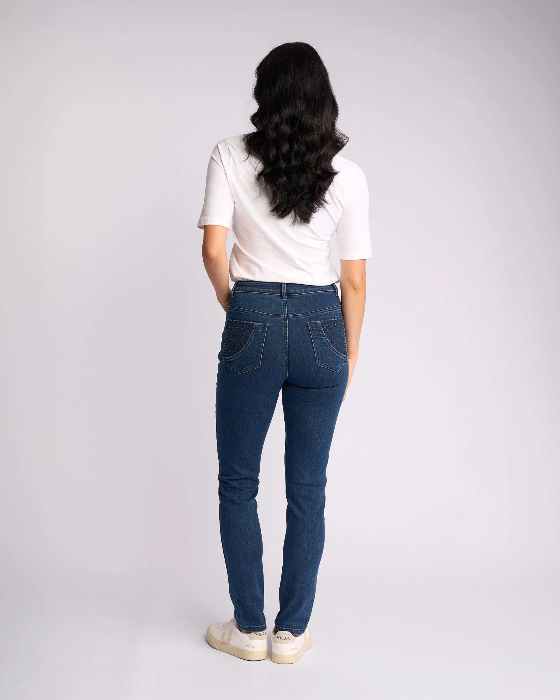 A person with long dark hair, wearing a white short-sleeve top, Vassalli Reign Jean - Brushed Indigo, and white sneakers stands facing away from the camera against a plain light background.