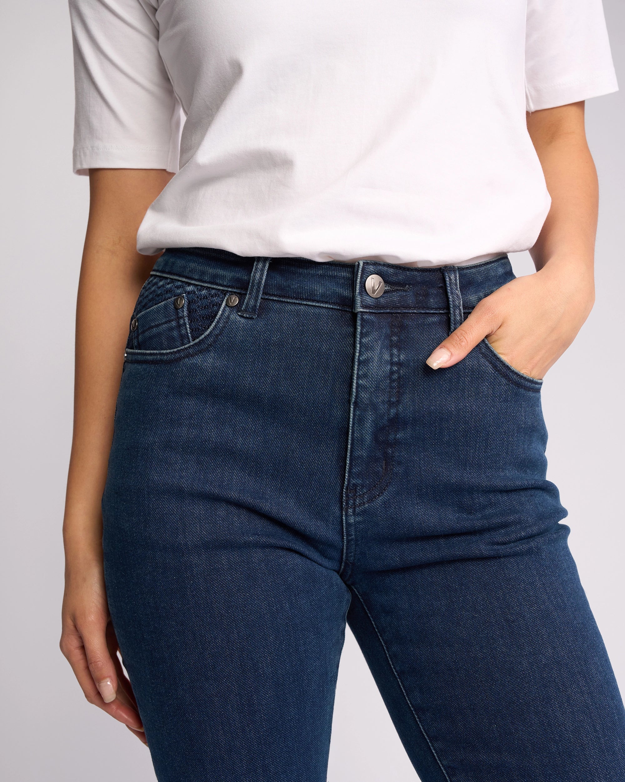 A person wearing a white short-sleeve shirt and Vassalli Reign Jean - Brushed Indigo skinny-leg jeans stands with one hand in their pocket against a plain light background.