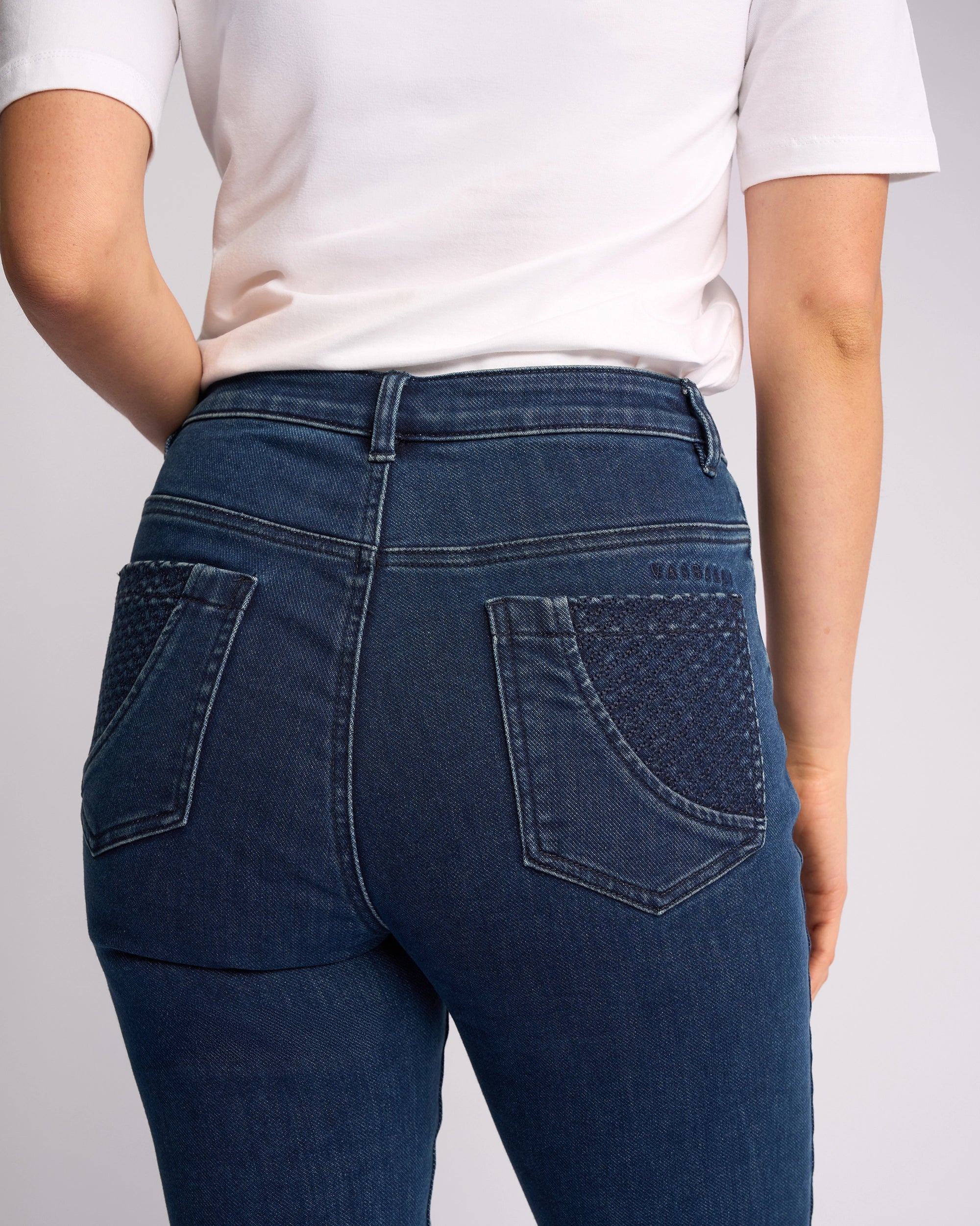 A person facing away from the camera wears a white short-sleeve shirt and Vassalli Reign Jean - Brushed Indigo, high-waisted blue skinny jeans with textured back pocket detailing.