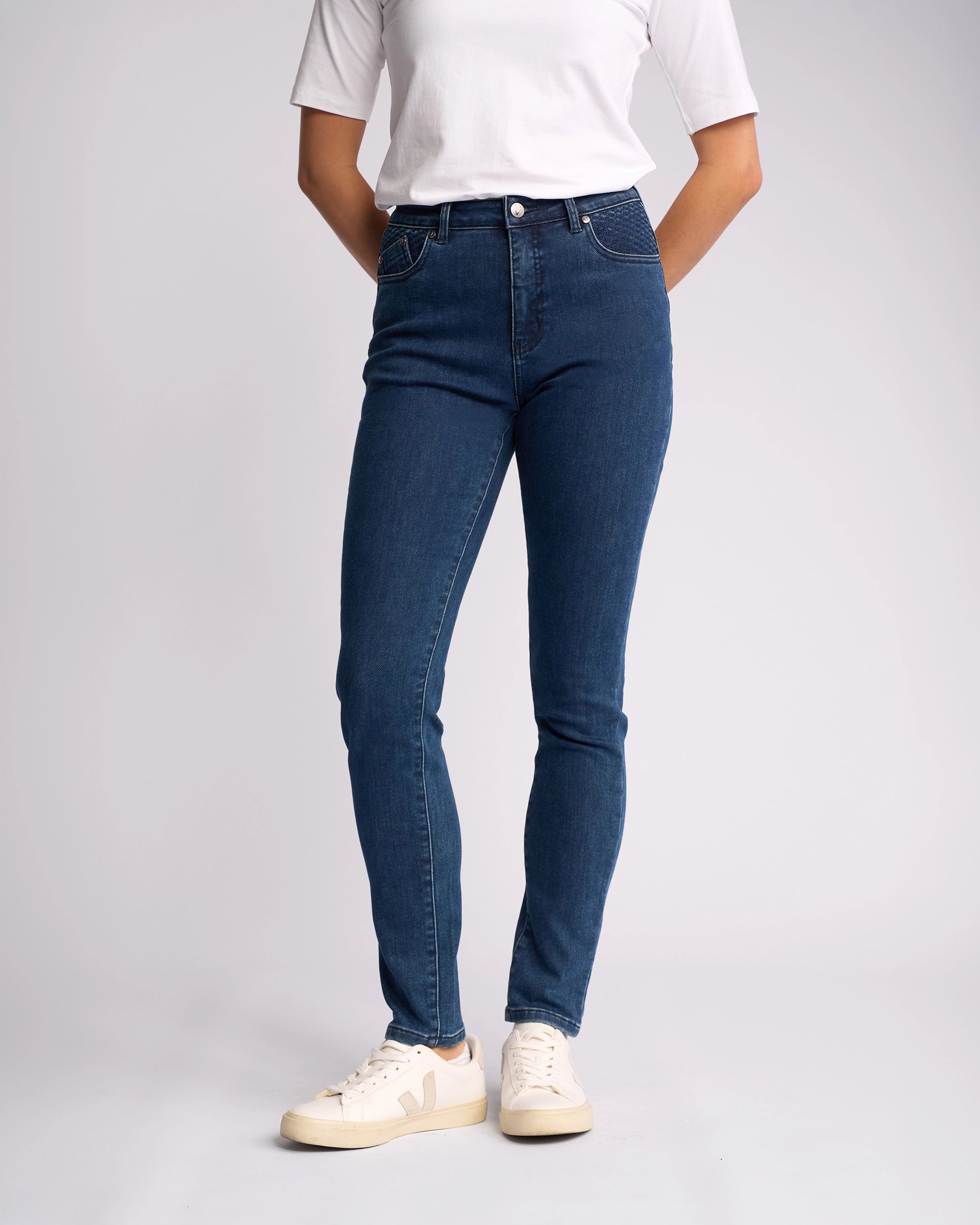 A person wearing a white short-sleeve shirt, Vassalli Reign Jean - Brushed Indigo, and white sneakers stands against a plain background with hands behind their back. Ideal for those seeking women’s denim NZ style.