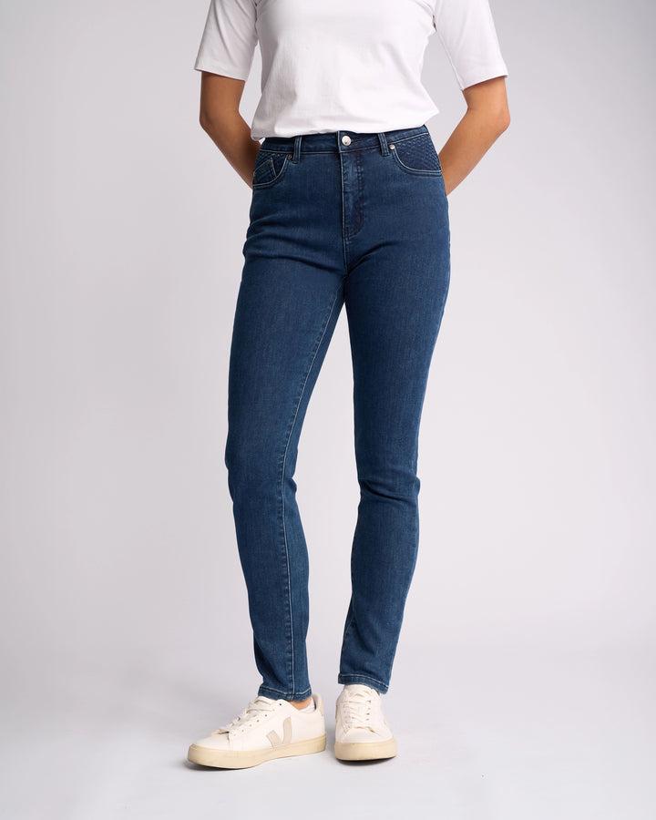 A person wearing a white short-sleeve shirt, Vassalli Reign Jean - Brushed Indigo, and white sneakers stands against a plain background with hands behind their back. Ideal for those seeking women’s denim NZ style.