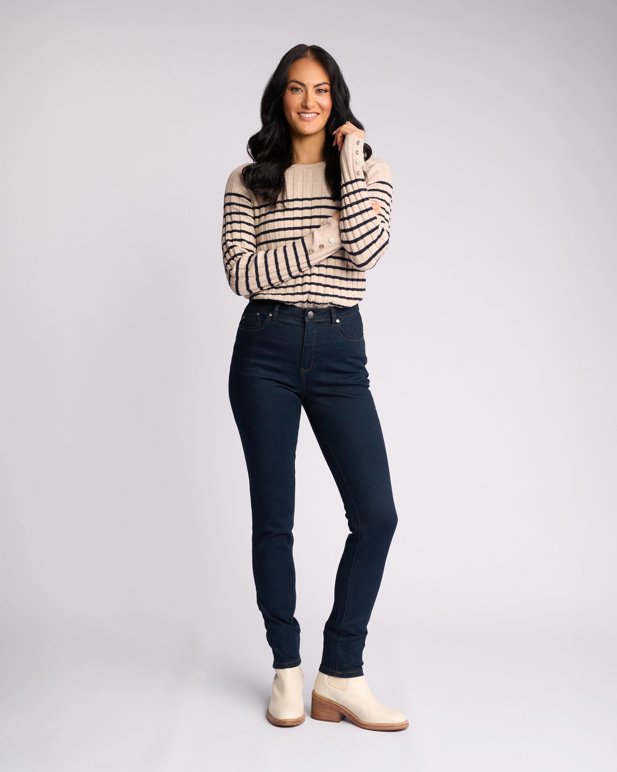 A woman with long dark hair smiles while standing, wearing the Vassalli Reign Jean in indigo denim, paired with a cream sweater with black stripes and beige ankle boots against a plain light background.