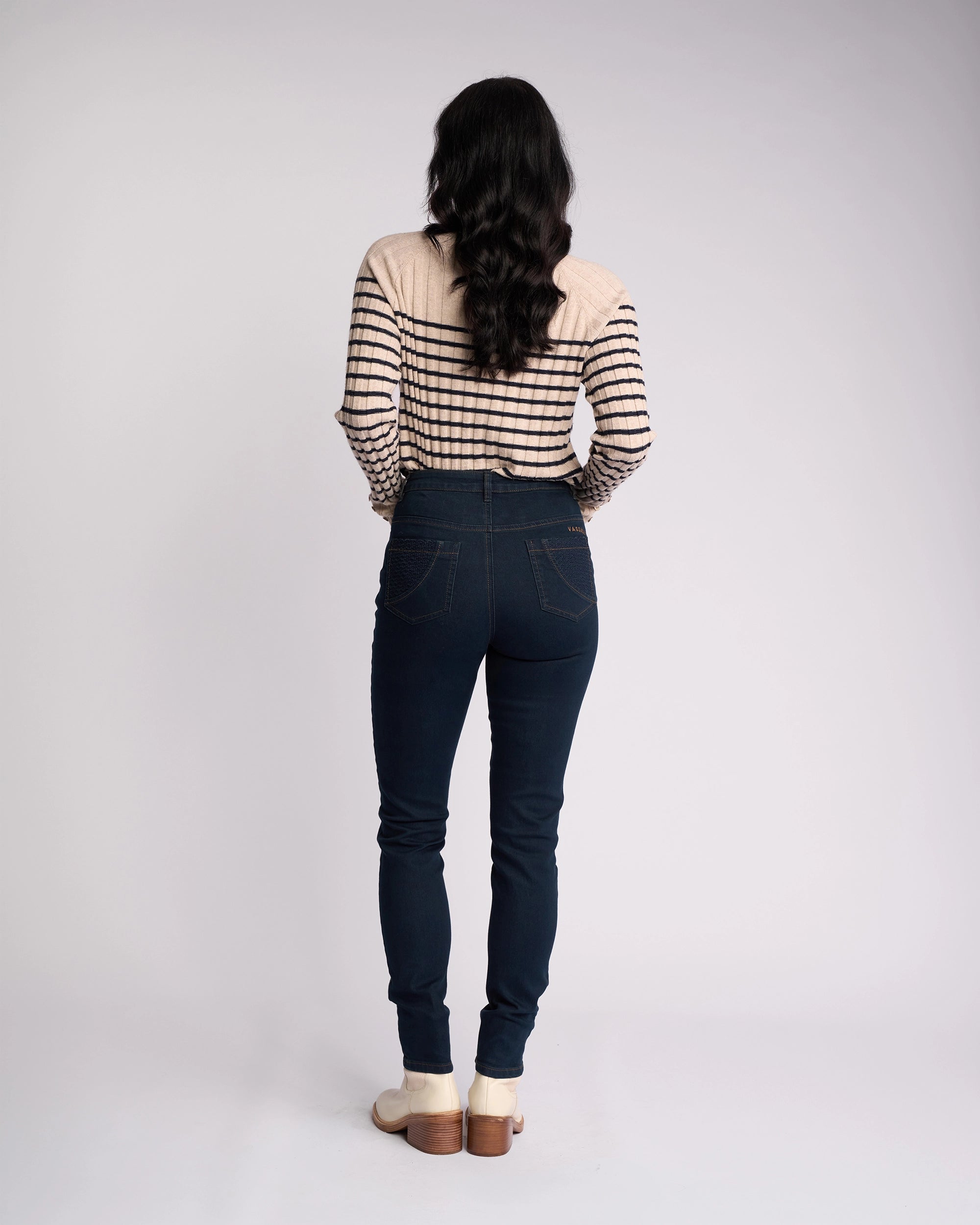 A person stands facing away, wearing indigo Reign Jean skinny-legs by Vassalli with a striped long-sleeve top and light shoes, set against a plain light background.