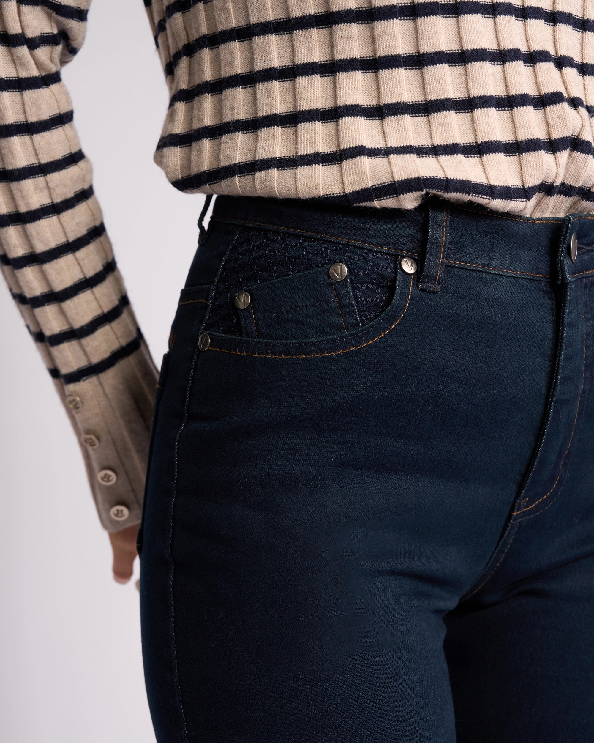 A person models the Vassalli Reign Jean - Indigo Denim, high-waisted skinny-leg style, paired with a beige and black striped long-sleeve shirt—showcasing classic women's NZ denim fashion from shoulders to mid-thigh.