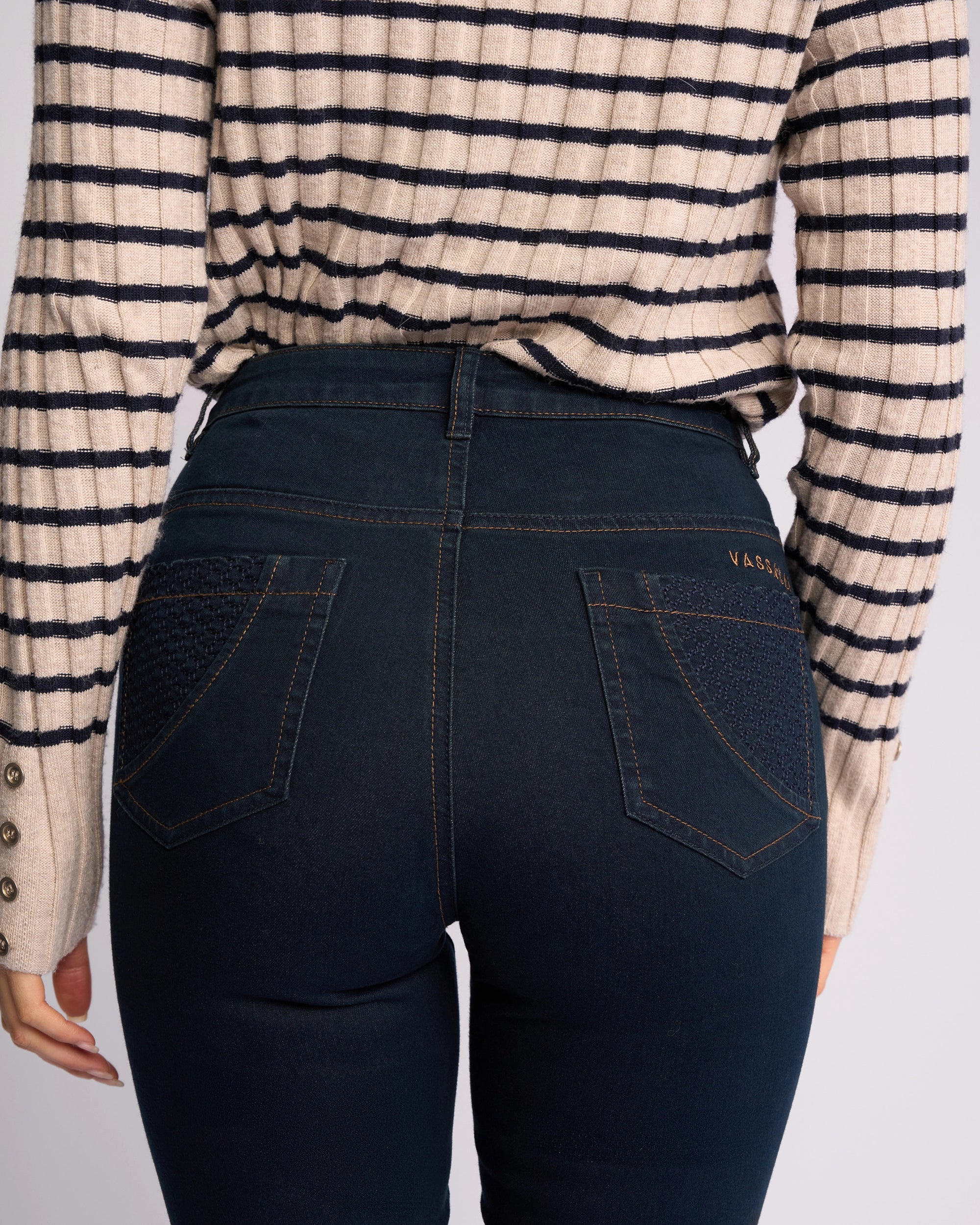 A woman, shown from the back, wears Vassalli's Reign Jean - Indigo Denim with detailed stitching and textured back pockets, paired with a cream, long-sleeve striped sweater featuring cuff buttons.