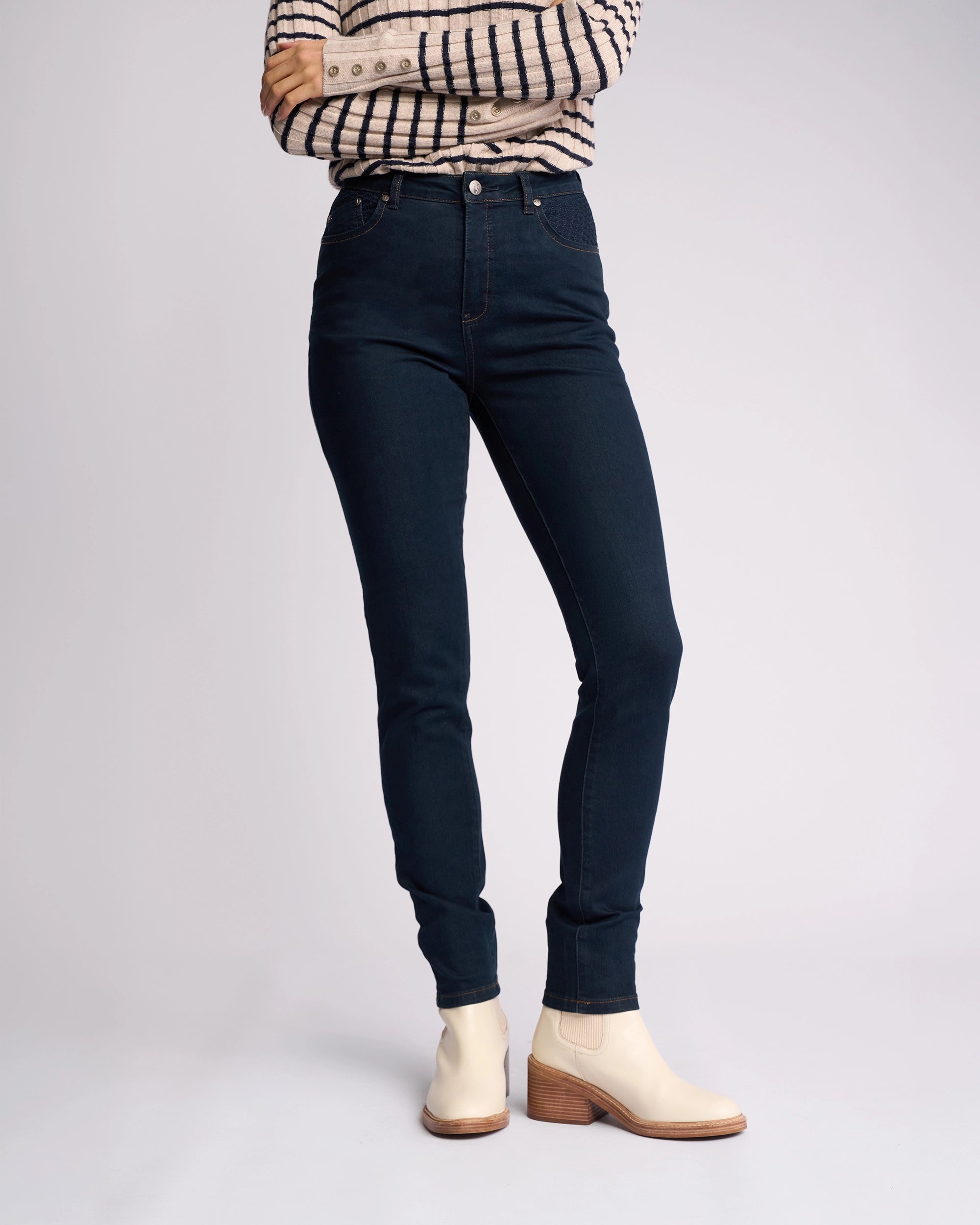 A person stands with arms crossed, wearing a plaid long-sleeve shirt, Vassalli Reign Jean in indigo denim, and beige ankle boots with wooden heels, against a plain light background.