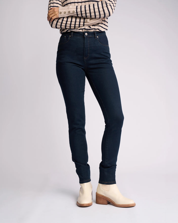 A person stands with arms crossed, wearing a plaid long-sleeve shirt, Vassalli Reign Jean in indigo denim, and beige ankle boots with wooden heels, against a plain light background.