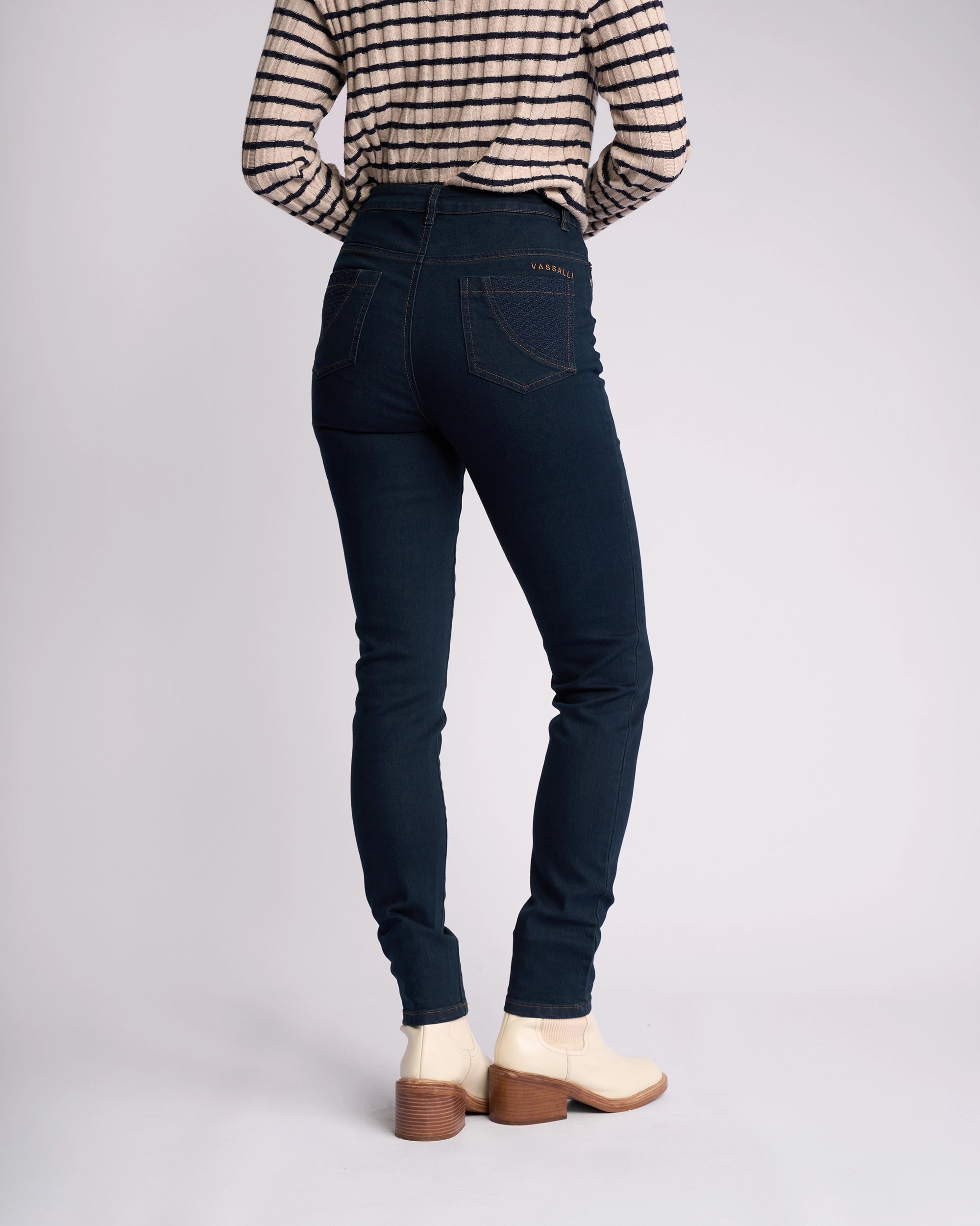 A person wearing Vassalli's Reign Jean in indigo denim, a black-and-white striped long-sleeve shirt, and white heeled ankle boots stands facing away against a plain background.
