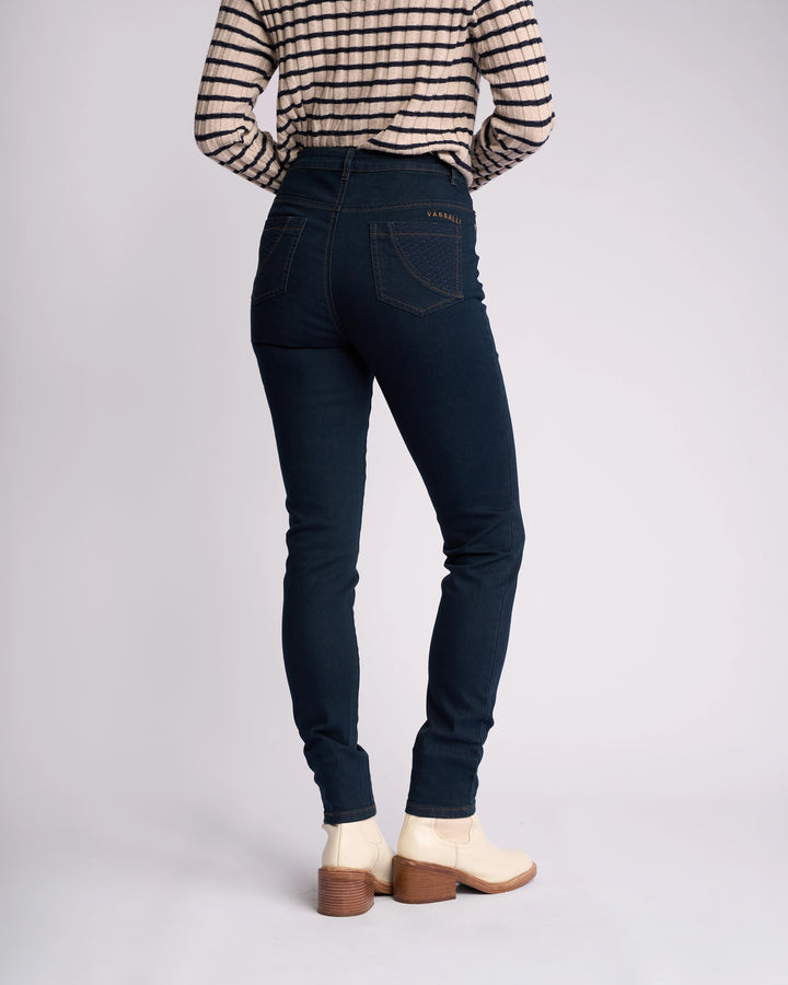 A person wearing Vassalli's Reign Jean in indigo denim, a black-and-white striped long-sleeve shirt, and white heeled ankle boots stands facing away against a plain background.