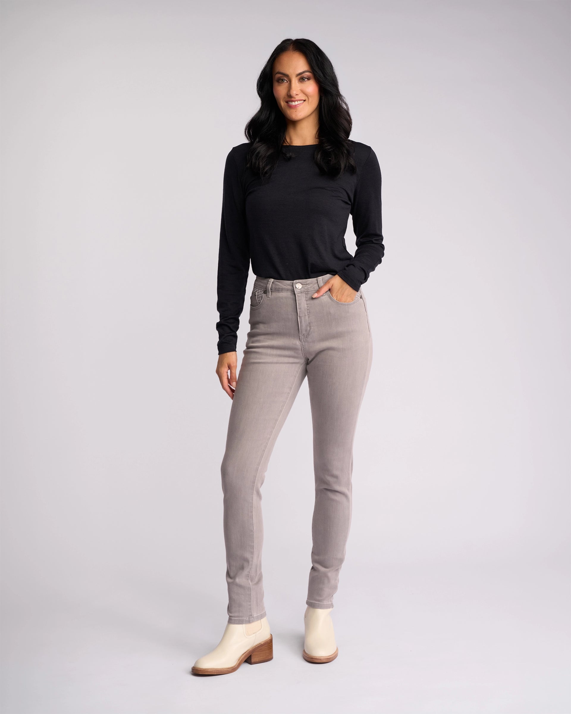 A woman with long dark hair wears a black long-sleeve top, Vassalli Reign Jean - Light Grey skinny-leg jeans, and off-white ankle boots, standing against a plain light gray background, smiling with one hand in her pocket.
