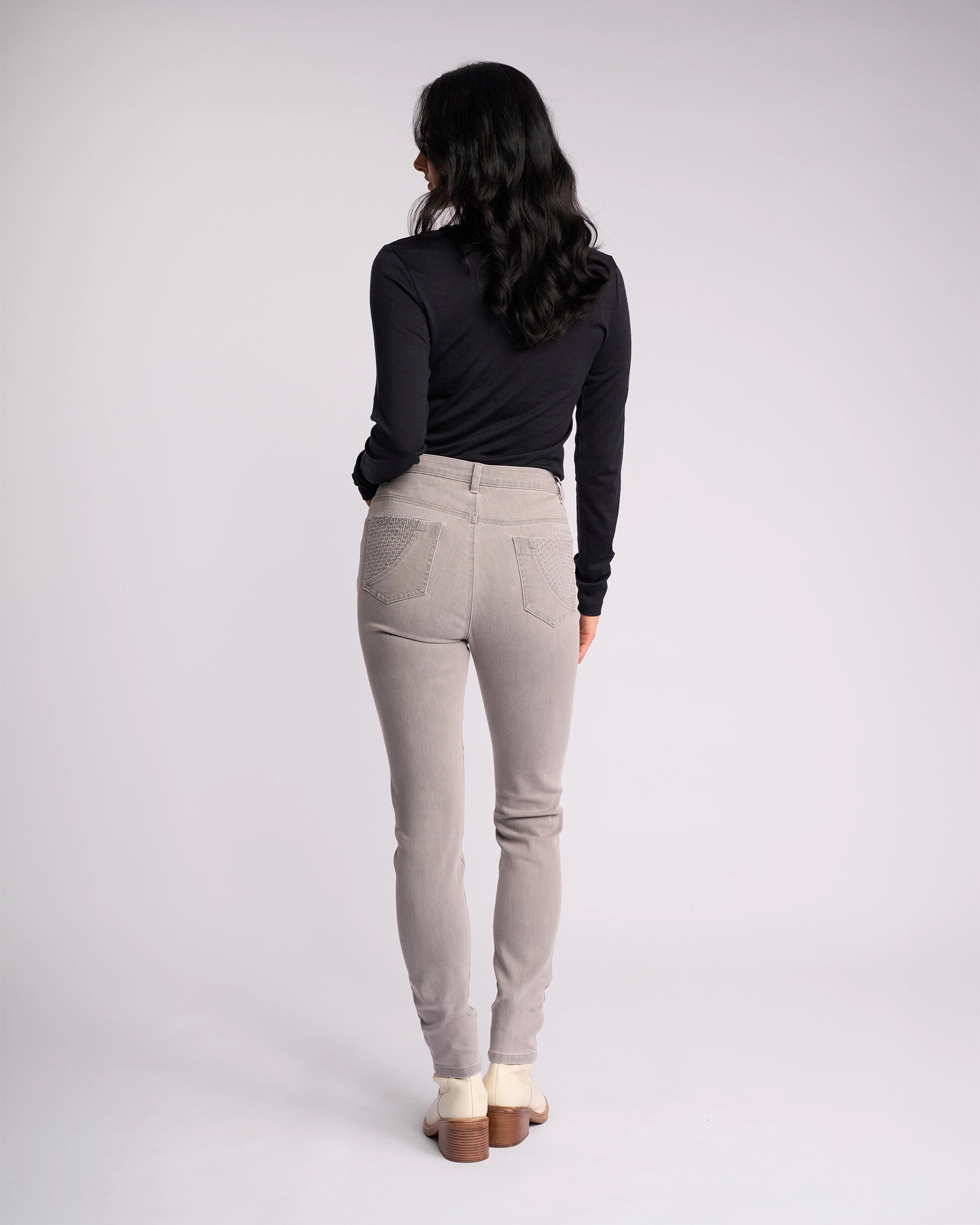 A woman with long dark hair faces away, wearing a black long-sleeve top, Vassalli Reign Jean - Light Grey, and cream ankle boots against a plain light background.