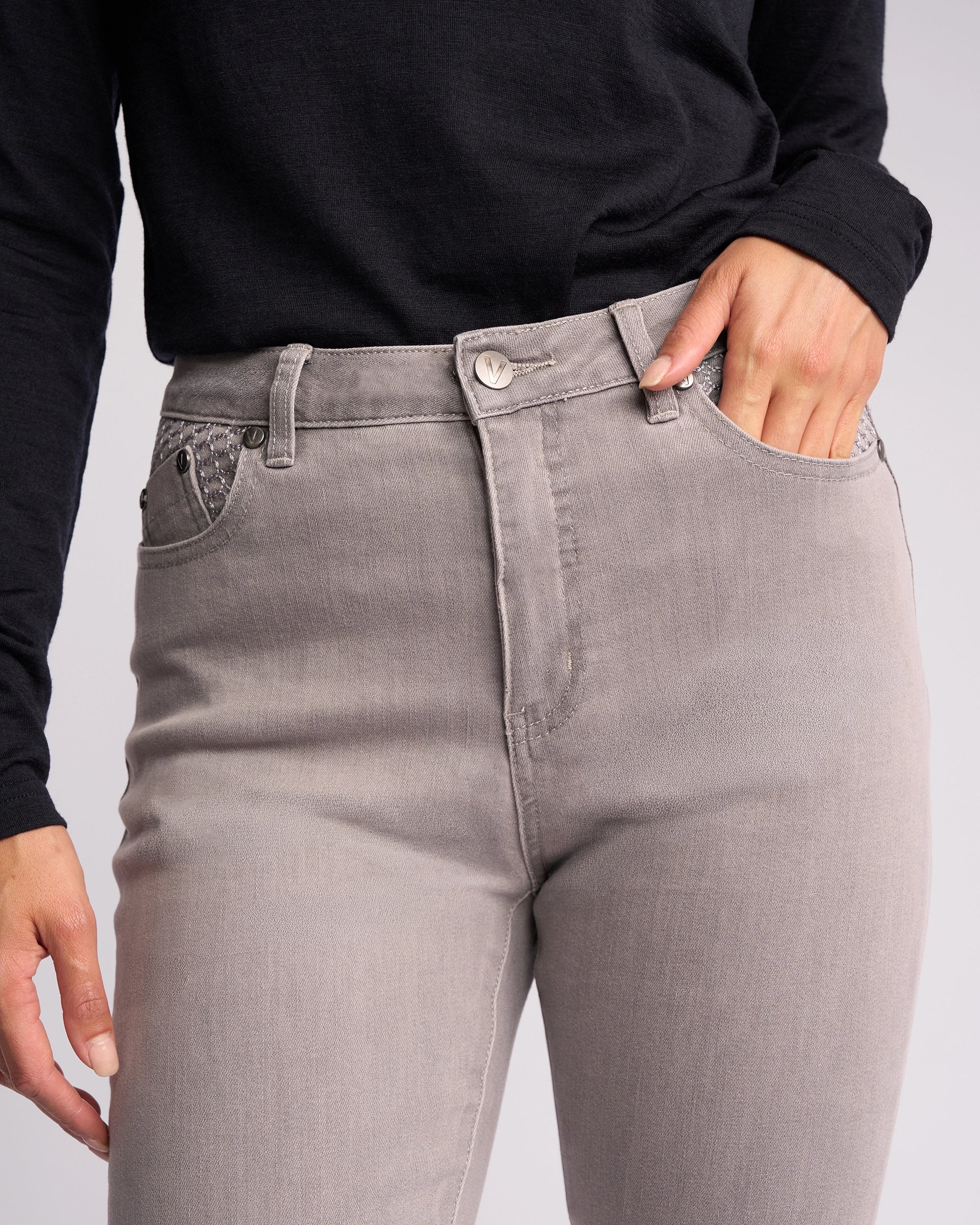 A person wears Vassalli Reign Jean - Light Grey, a high-waisted skinny-leg women’s denim, paired with a black long-sleeve shirt and one hand in the front pocket.