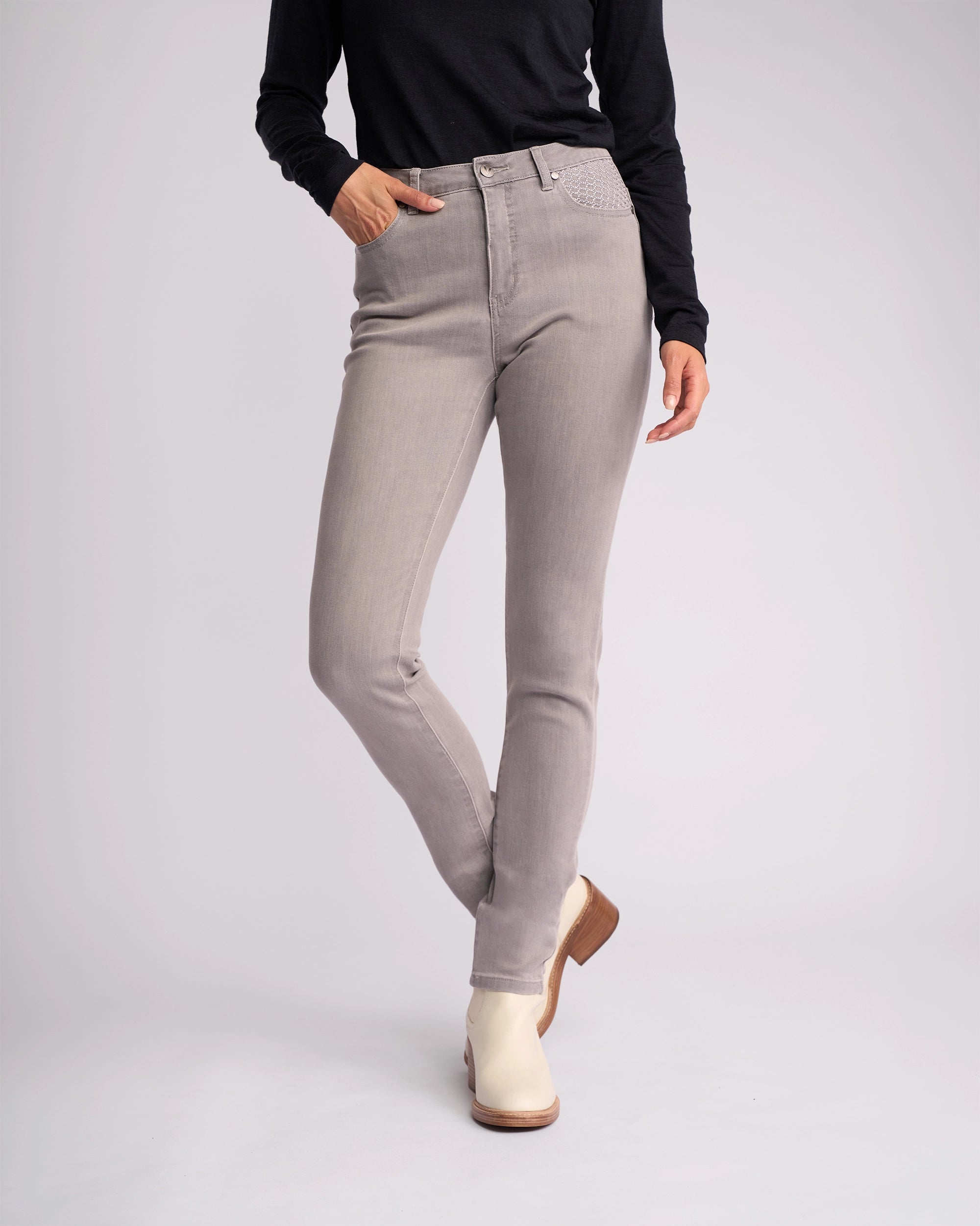 A person models the Vassalli Reign Jean - Light Grey, paired with a black long-sleeve top and off-white ankle boots, standing against a plain white background with one hand in their pocket.