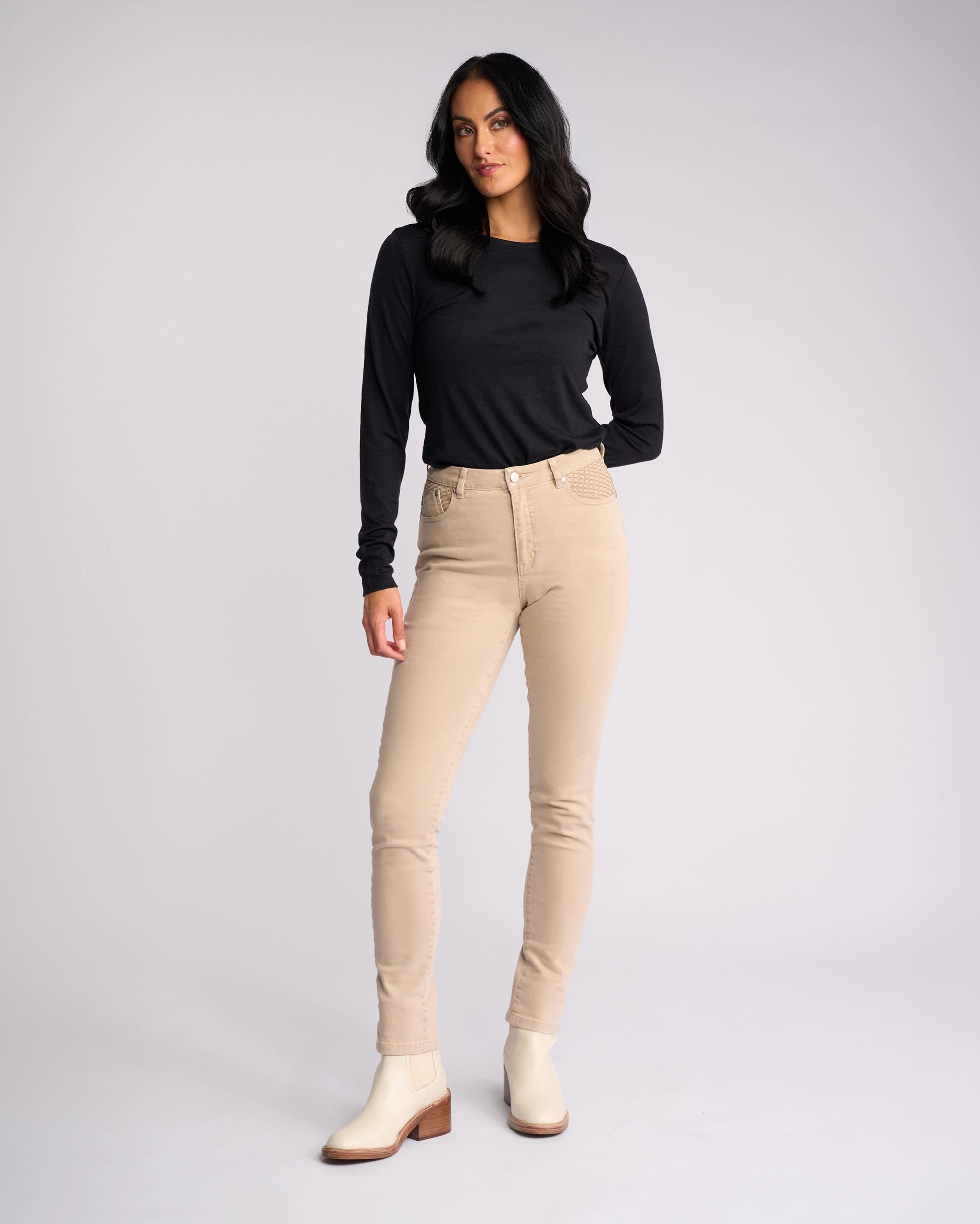 A woman with long dark hair wears a black top, off-white ankle boots, and the Vassalli Reign Jean in oat denim, standing against a plain light background.