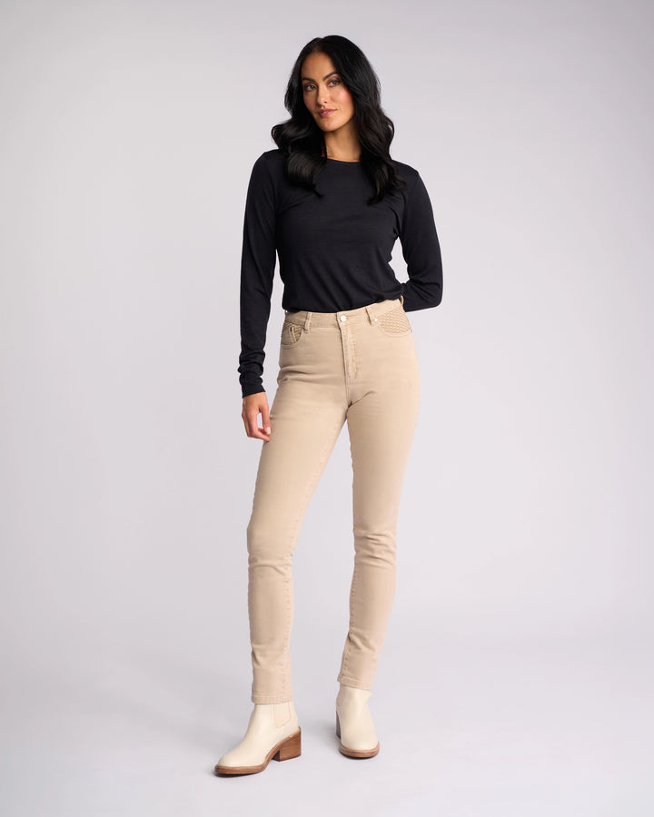 A woman with long dark hair wears a black top, off-white ankle boots, and the Vassalli Reign Jean in oat denim, standing against a plain light background.