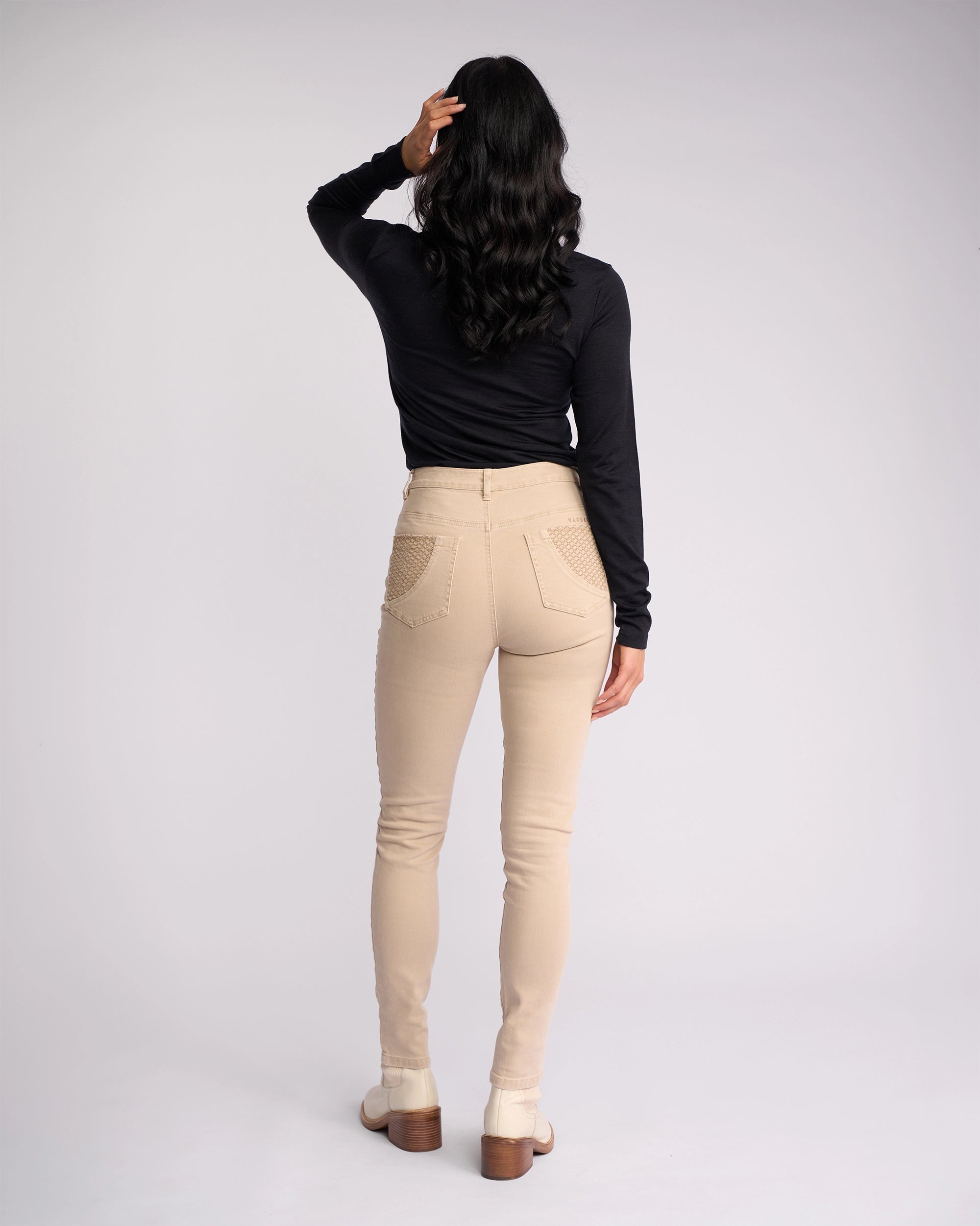 A woman stands with her back to the camera, wearing a black long-sleeve top and Vassalli's Reign Jean in Oat Denim. One arm is raised to touch her hair, against a plain light background—perfect for fans of women’s denim NZ style.