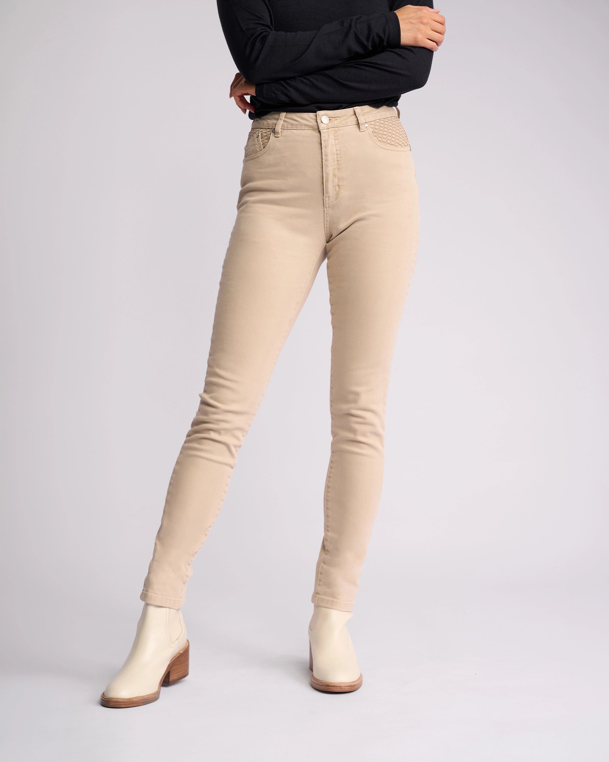 A person stands with arms crossed against a light grey background, wearing Vassalli Reign Jean - Oat Denim high-waisted skinny jeans, a black long-sleeve top, and cream ankle boots. Their head is not visible.