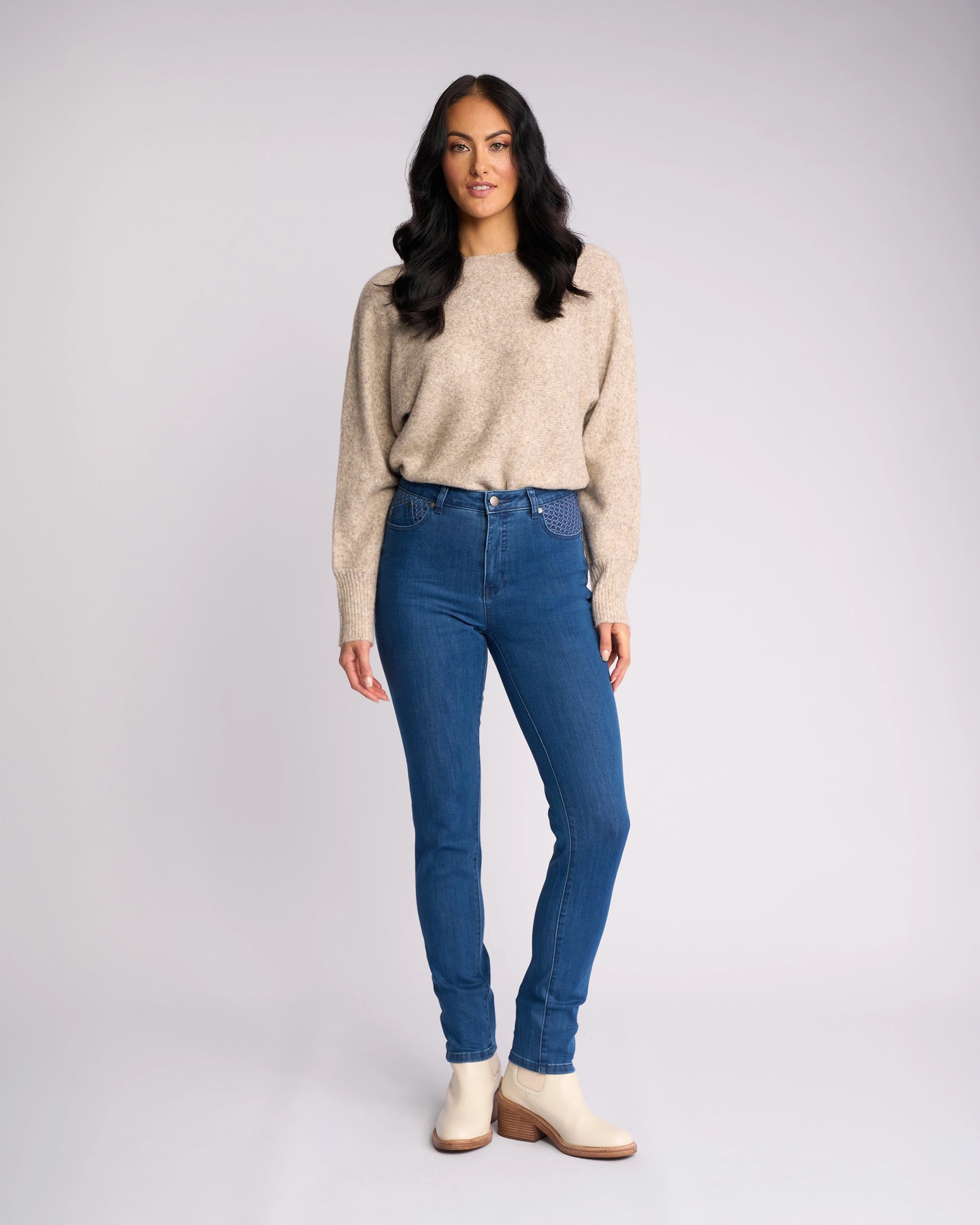 A woman with long, wavy dark hair stands against a plain light background, wearing Vassalli's Reign Jean - Plain New Blue, a beige sweater, and light ankle boots. She looks at the camera with a neutral expression.
