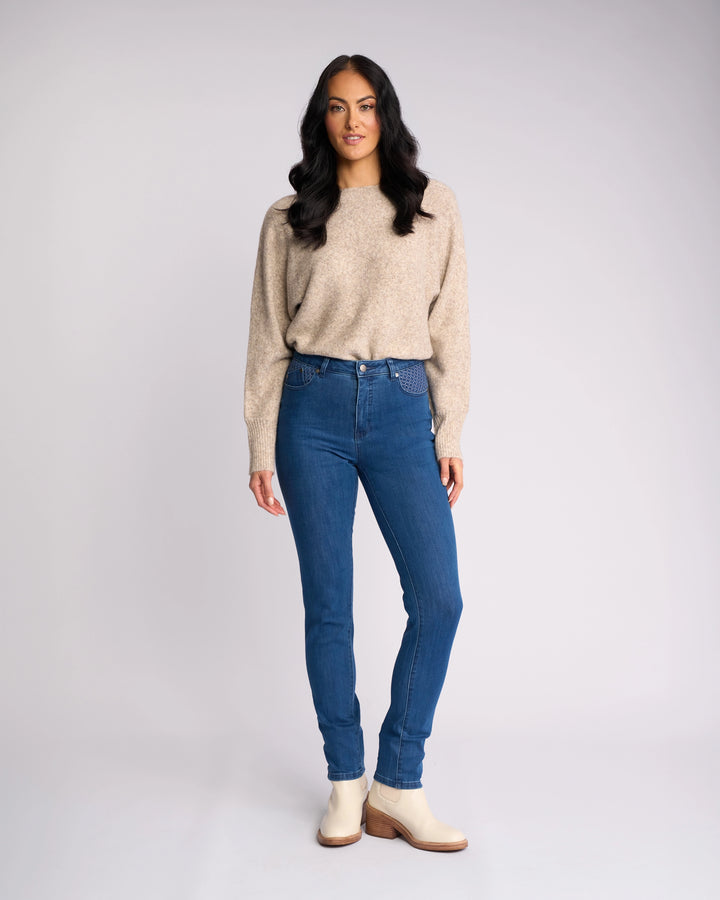 A woman with long, wavy dark hair stands against a plain light background, wearing Vassalli's Reign Jean - Plain New Blue, a beige sweater, and light ankle boots. She looks at the camera with a neutral expression.