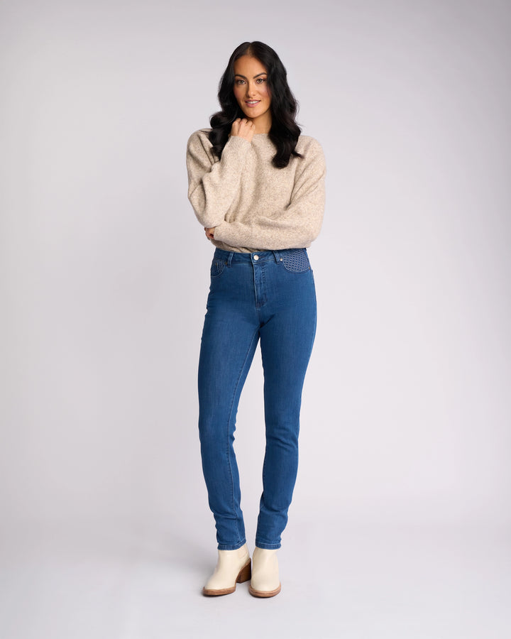 A woman with long dark hair poses against a plain light background, wearing a beige sweater, Vassalli Reign Jean - Plain New Blue, and white ankle boots. She smiles slightly, one hand touching her chin.