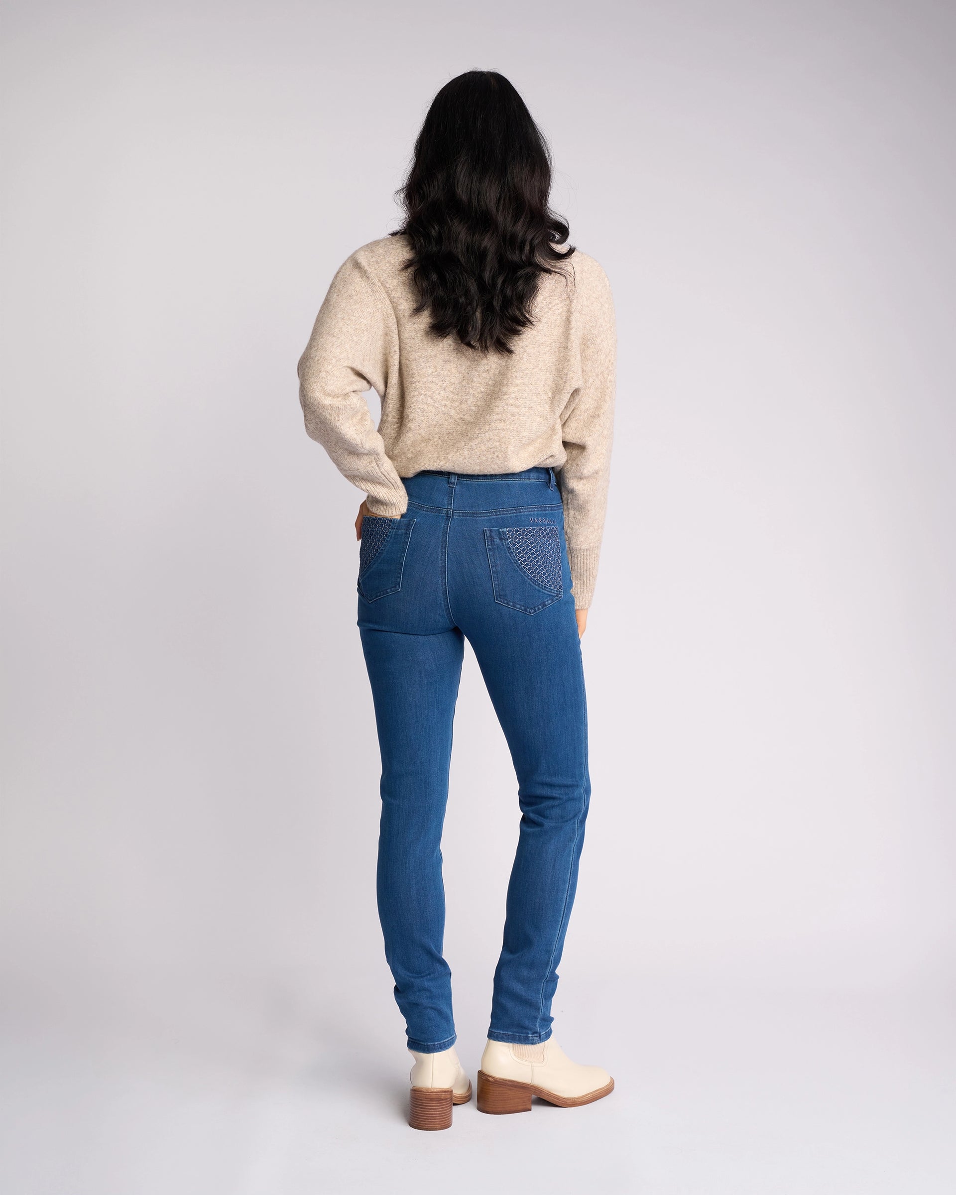 A person with long dark hair faces away, wearing a beige sweater, Vassalli Reign Jean - Plain New Blue from the women's denim NZ collection, and cream ankle boots against a plain light background.