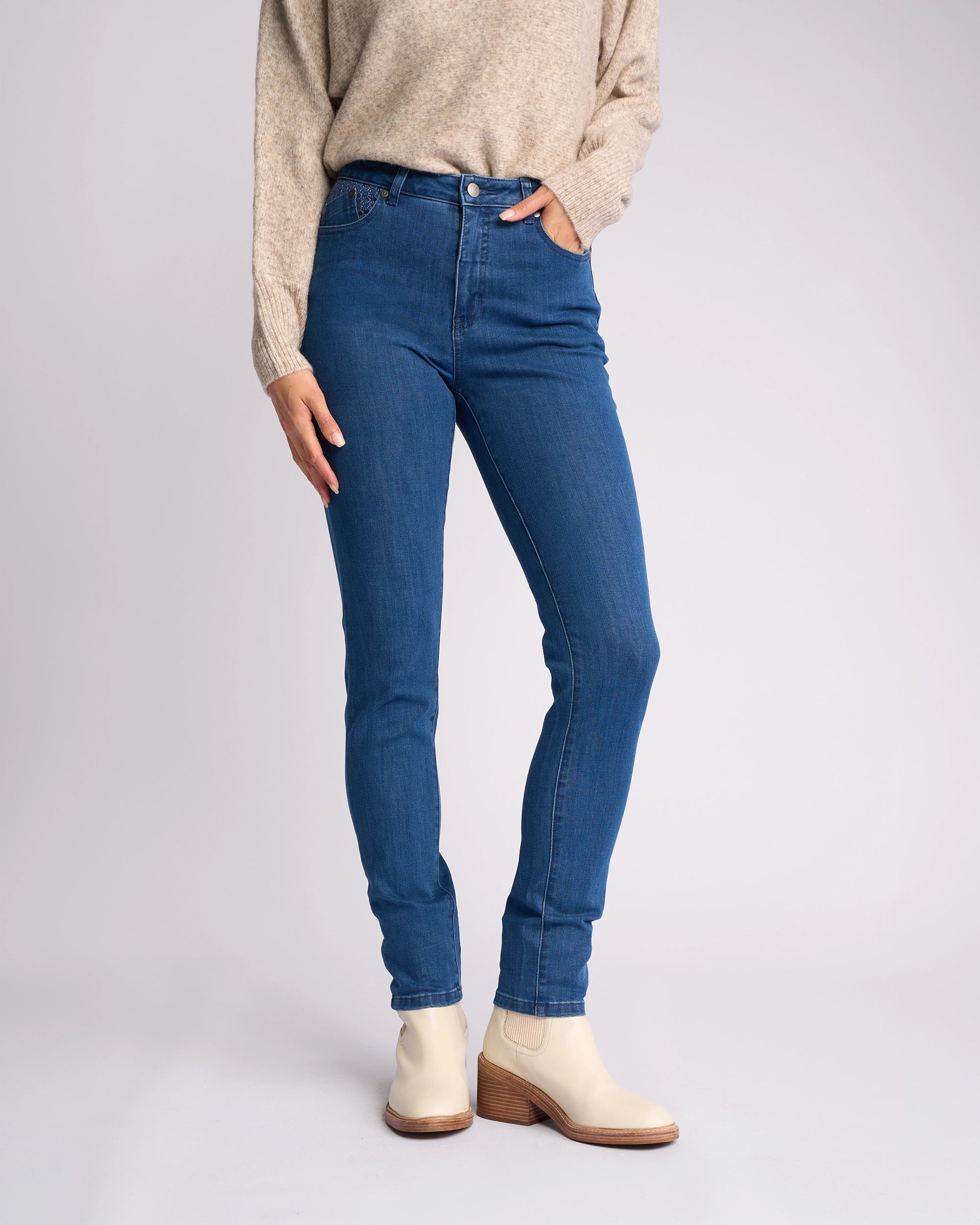 A person wearing a beige sweater, Vassalli Reign Jean - Plain New Blue skinny-leg jeans, and off-white ankle boots with wooden heels stands against a plain light background.