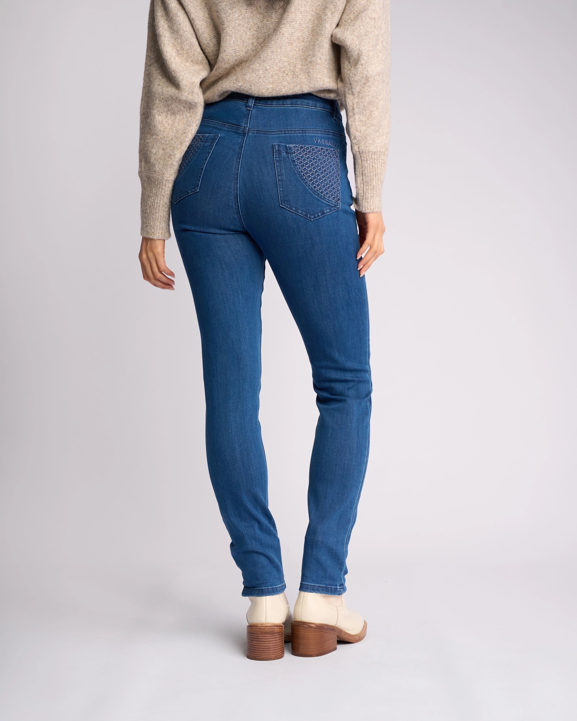 A person is shown from the back wearing a beige sweater, Vassalli Reign Jean - Plain New Blue, and light-colored ankle boots with wooden heels, standing against a plain light background.
