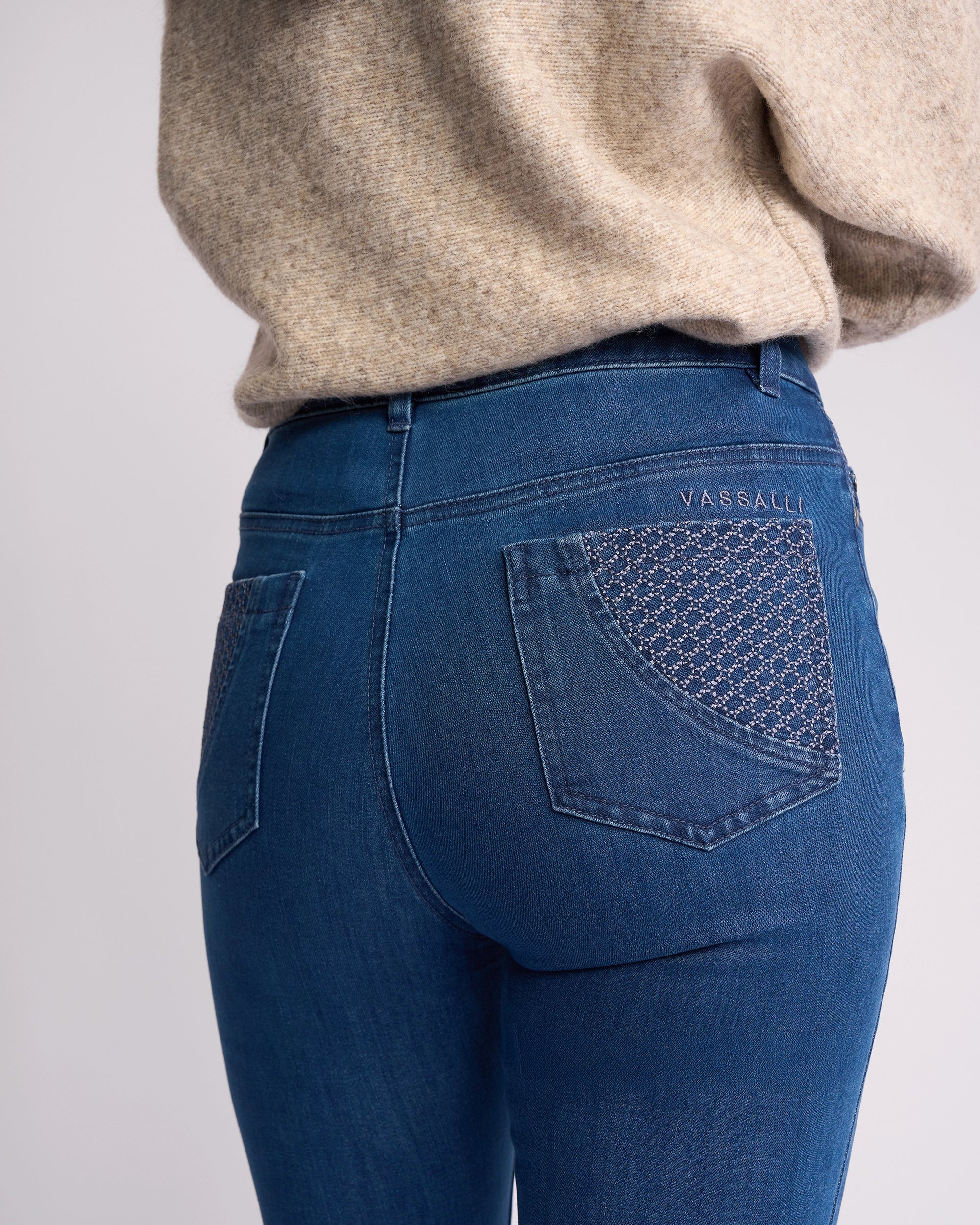 A person wears Vassalli’s Reign Jean - Plain New Blue skinny-leg women’s denim with plain back pockets, paired with a beige knit sweater. "Vassalli" is embroidered above the right pocket; shown from the waist down and back.