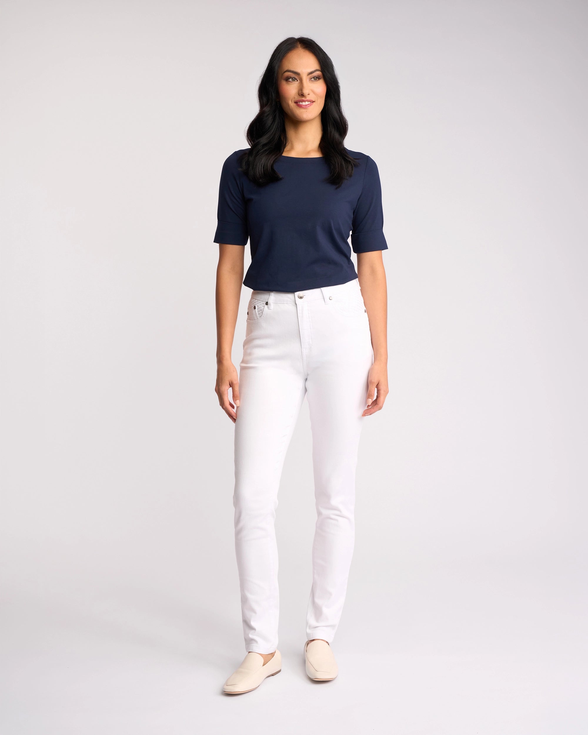 A woman with long dark hair stands and smiles, wearing a navy blue short-sleeve top, Vassalli's Reign Jean in White Denim, and light shoes against a plain light background.