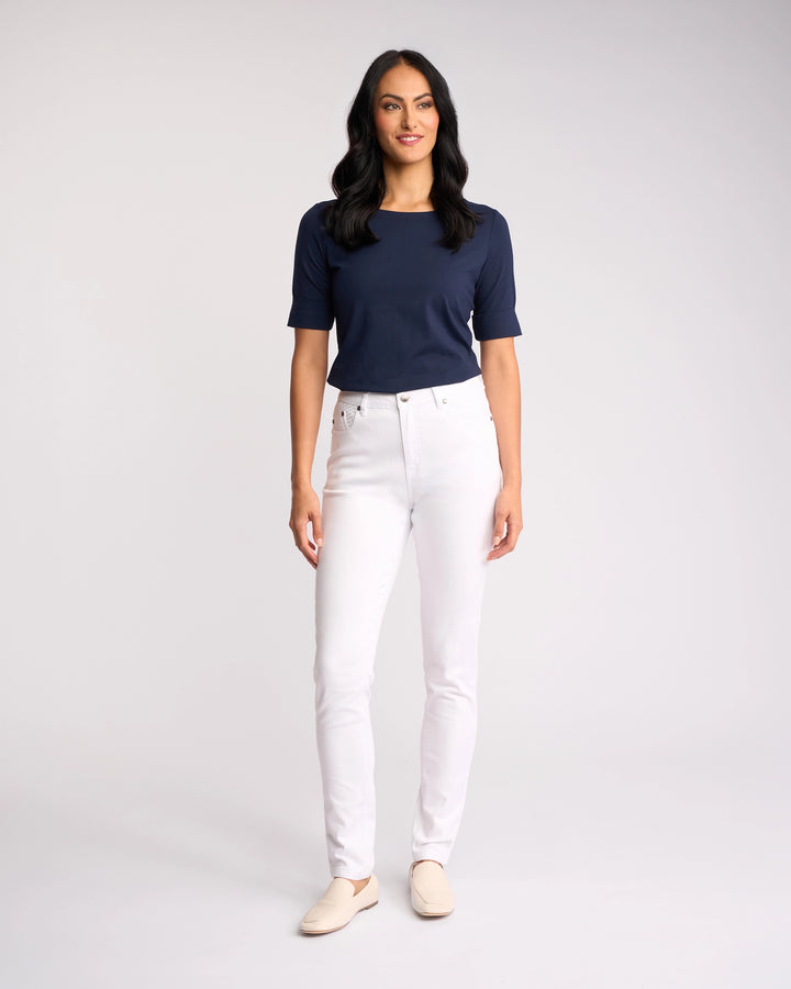 A woman with long dark hair stands and smiles, wearing a navy blue short-sleeve top, Vassalli's Reign Jean in White Denim, and light shoes against a plain light background.