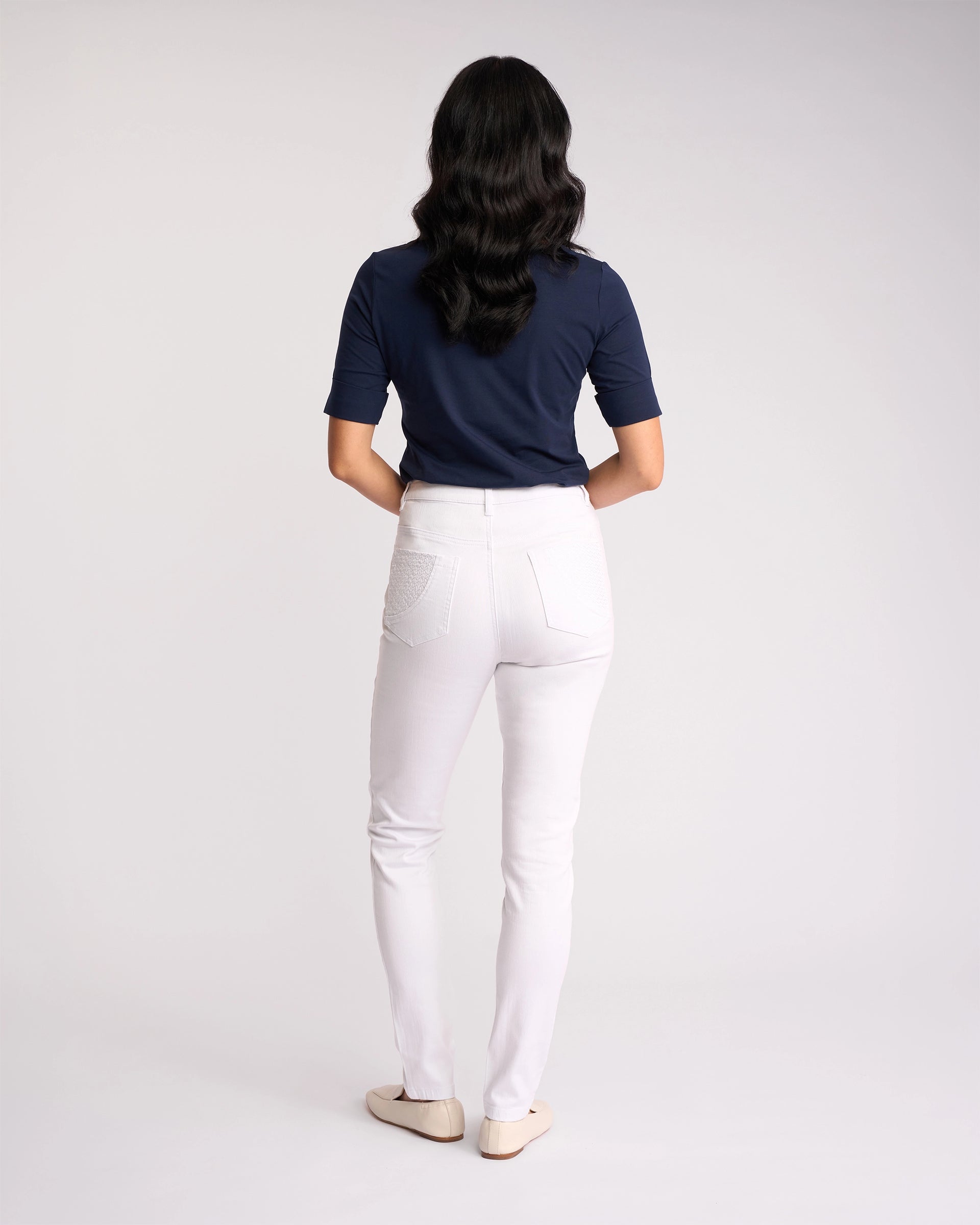 A person with long wavy hair, facing away from the camera, wears a navy short-sleeve top, Vassalli Reign Jean - White Denim, and beige flats against a plain white background.