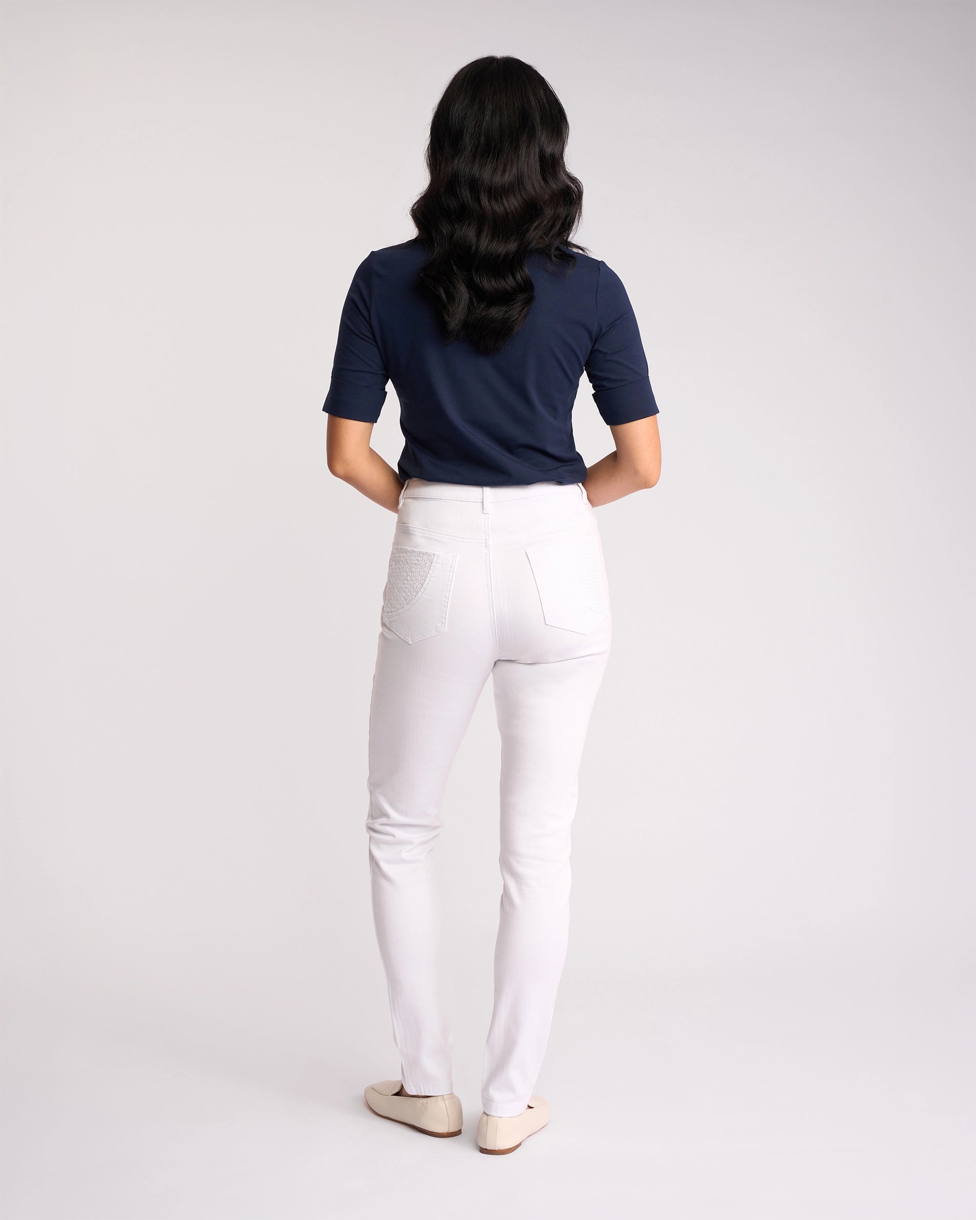A person with long wavy hair, facing away from the camera, wears a navy short-sleeve top, Vassalli Reign Jean - White Denim, and beige flats against a plain white background.
