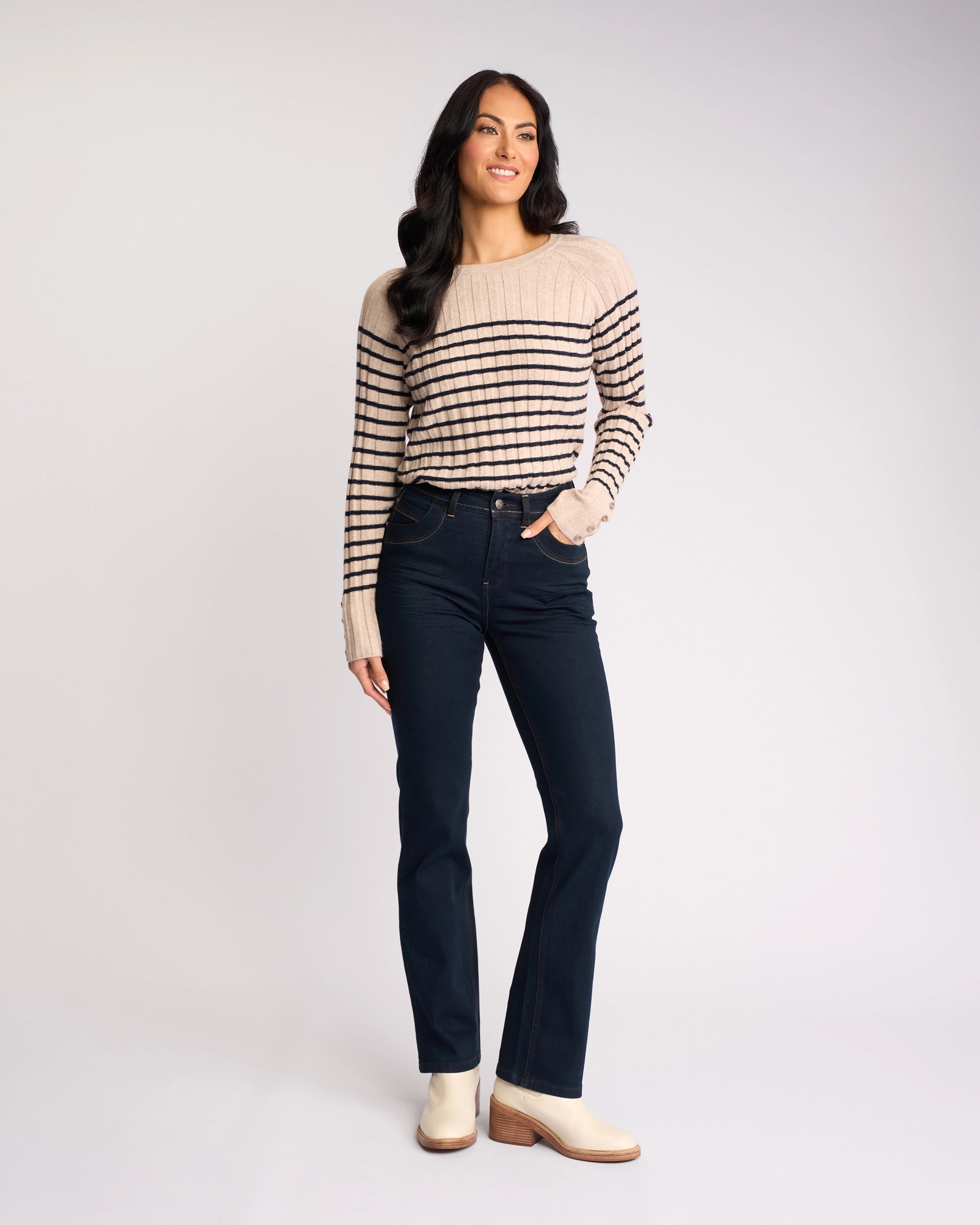 A woman with long dark hair wears the Vassalli Pace Jean in indigo denim, paired with a beige and black striped sweater and light ankle boots. She stands smiling with one hand in her pocket against a plain light background.