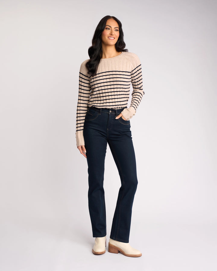 A woman with long dark hair wears the Vassalli Pace Jean in indigo denim, paired with a beige and black striped sweater and light ankle boots. She stands smiling with one hand in her pocket against a plain light background.