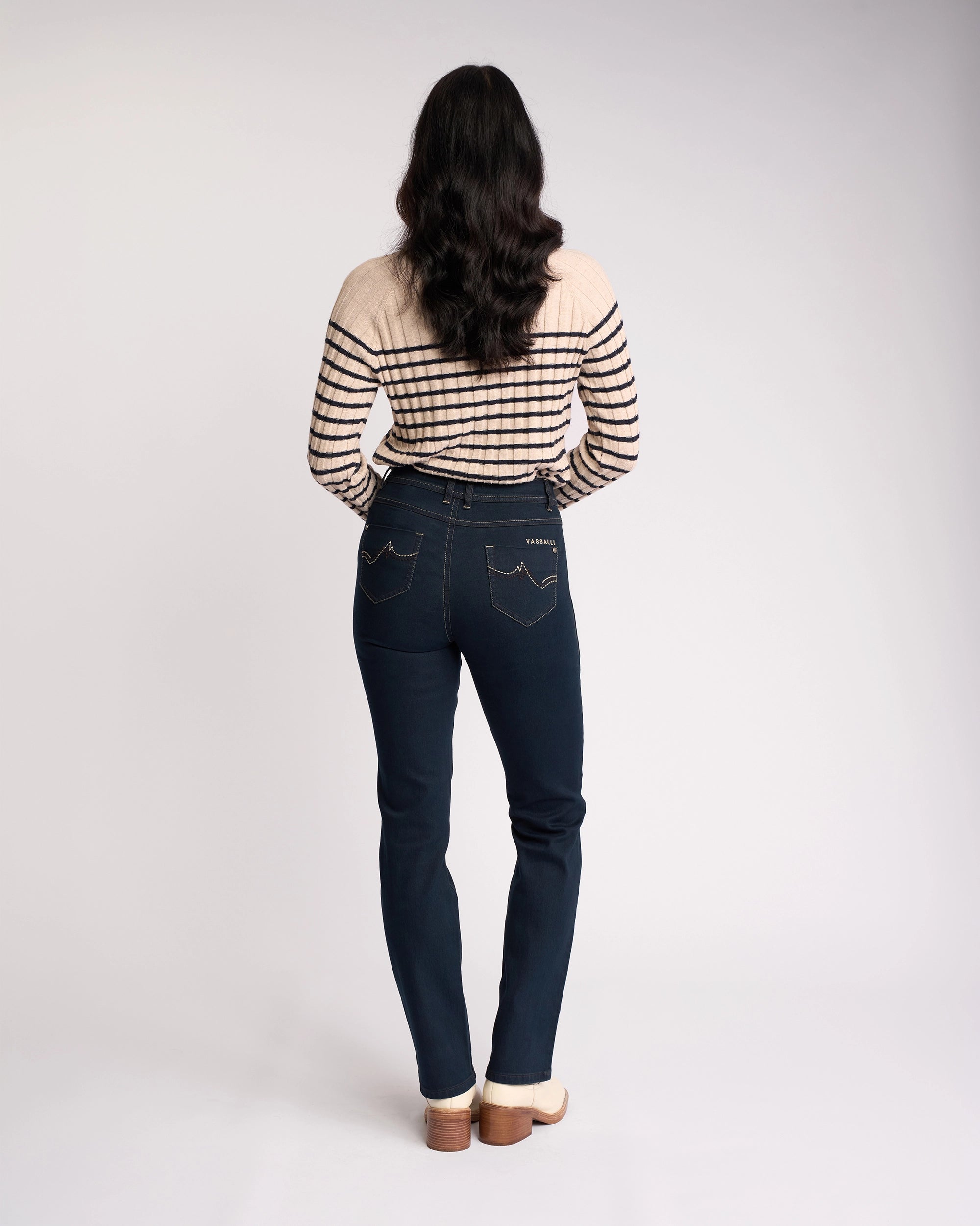 A person with long wavy hair, dressed in a beige and black striped sweater, Vassalli's Pace Jean in Indigo Denim, and light shoes stands with their back to the camera against a plain light background.
