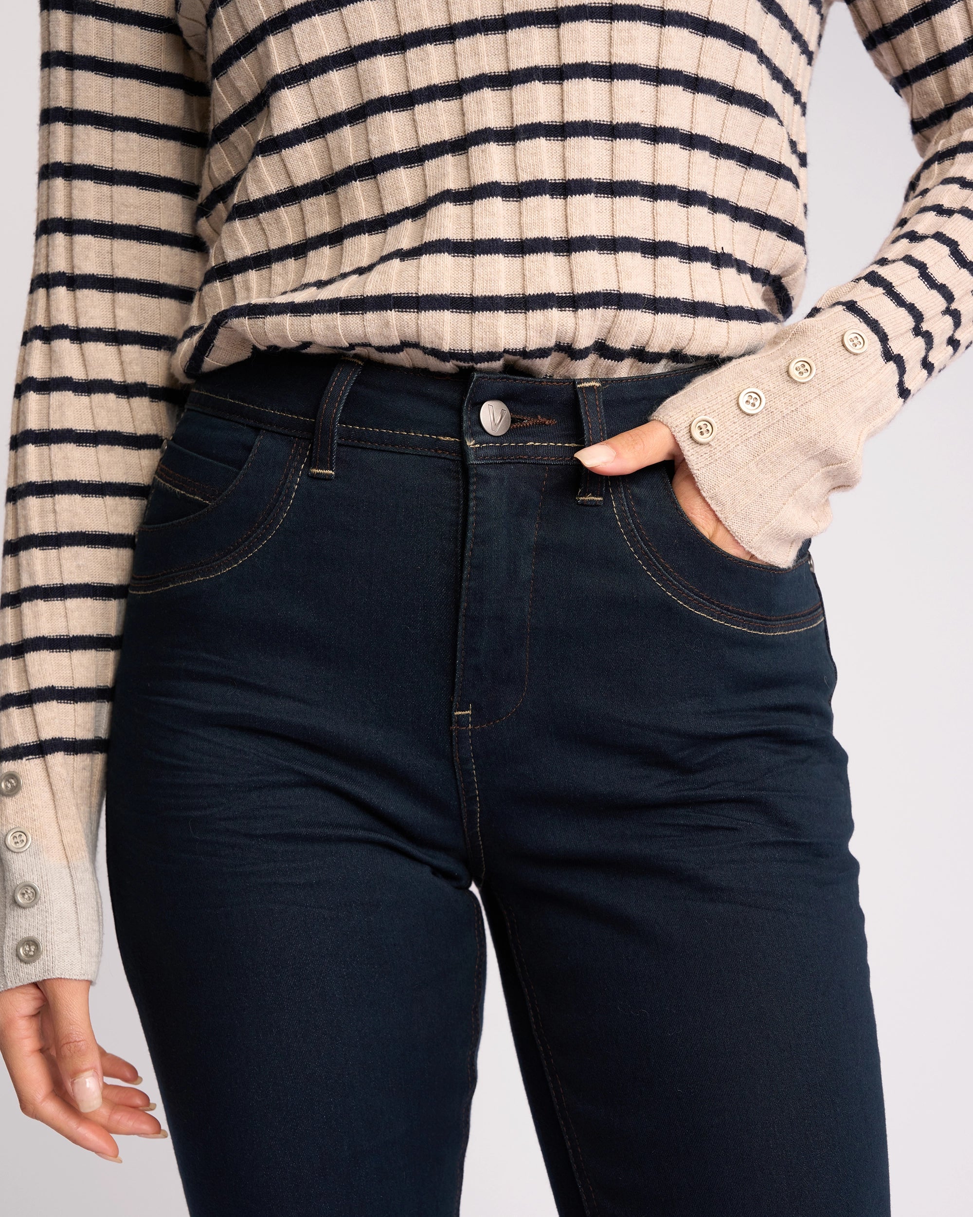 A person wears Vassalli’s Pace Jean in indigo denim, paired with a beige and black striped long-sleeve top featuring cuff buttons, and has one hand in a pocket.